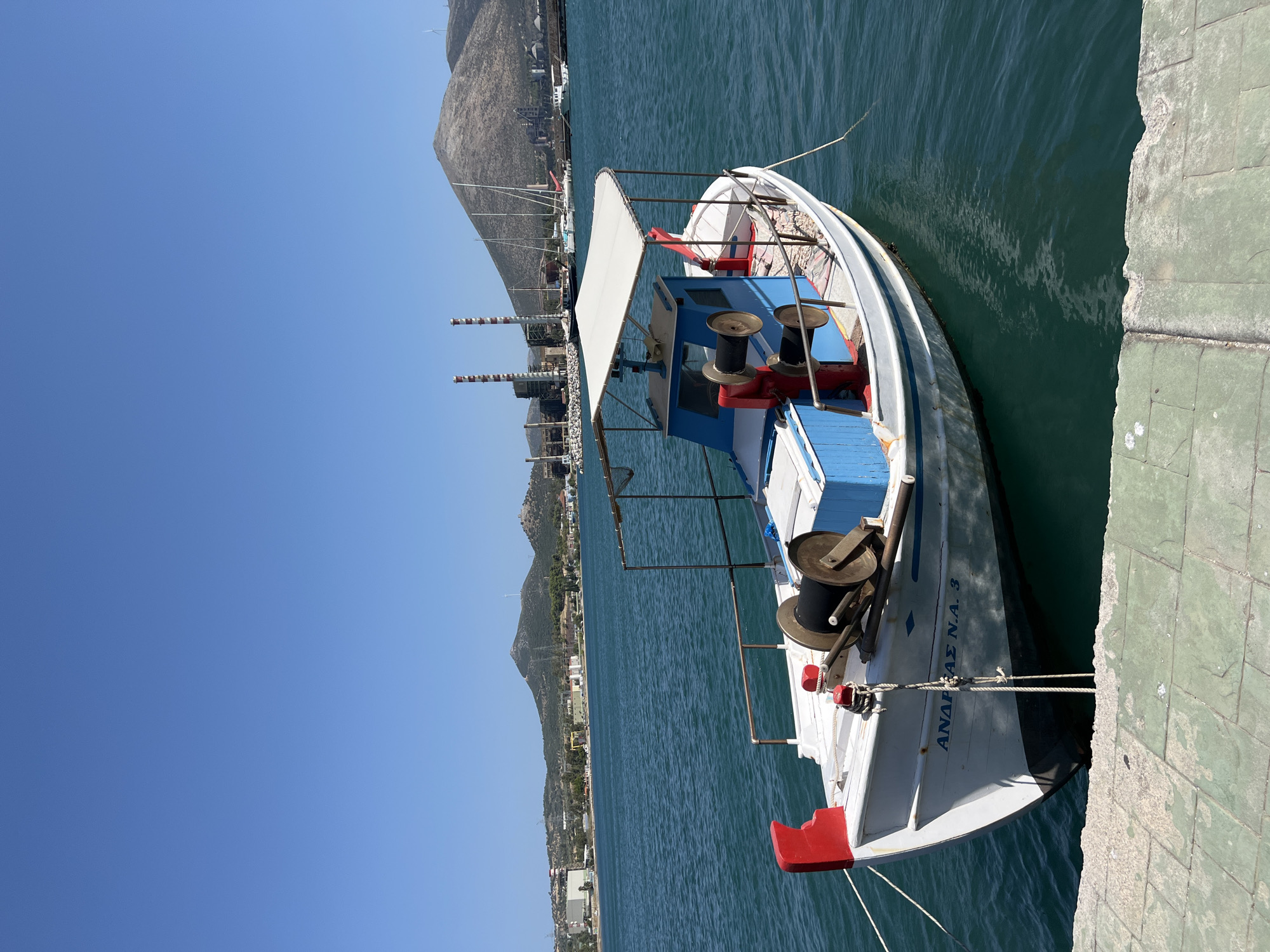 A white and blue fishing boat is moored in the turquoise water of a harbor in Karavos, Greece, on a clear, sunny day. The boat has a white canopy over a blue cabin. Red and black winches are visible on the deck. The name "ΑΝΔΡΕΑΣ Ν.Α. 3" is painted on the hull. In the background, a line of industrial buildings, including smokestacks, is visible on the shore, along with a large, barren mountain and a few sailboats. The water is calm, with a slight ripple. A concrete walkway is in the foreground. There are no people visible in the image.