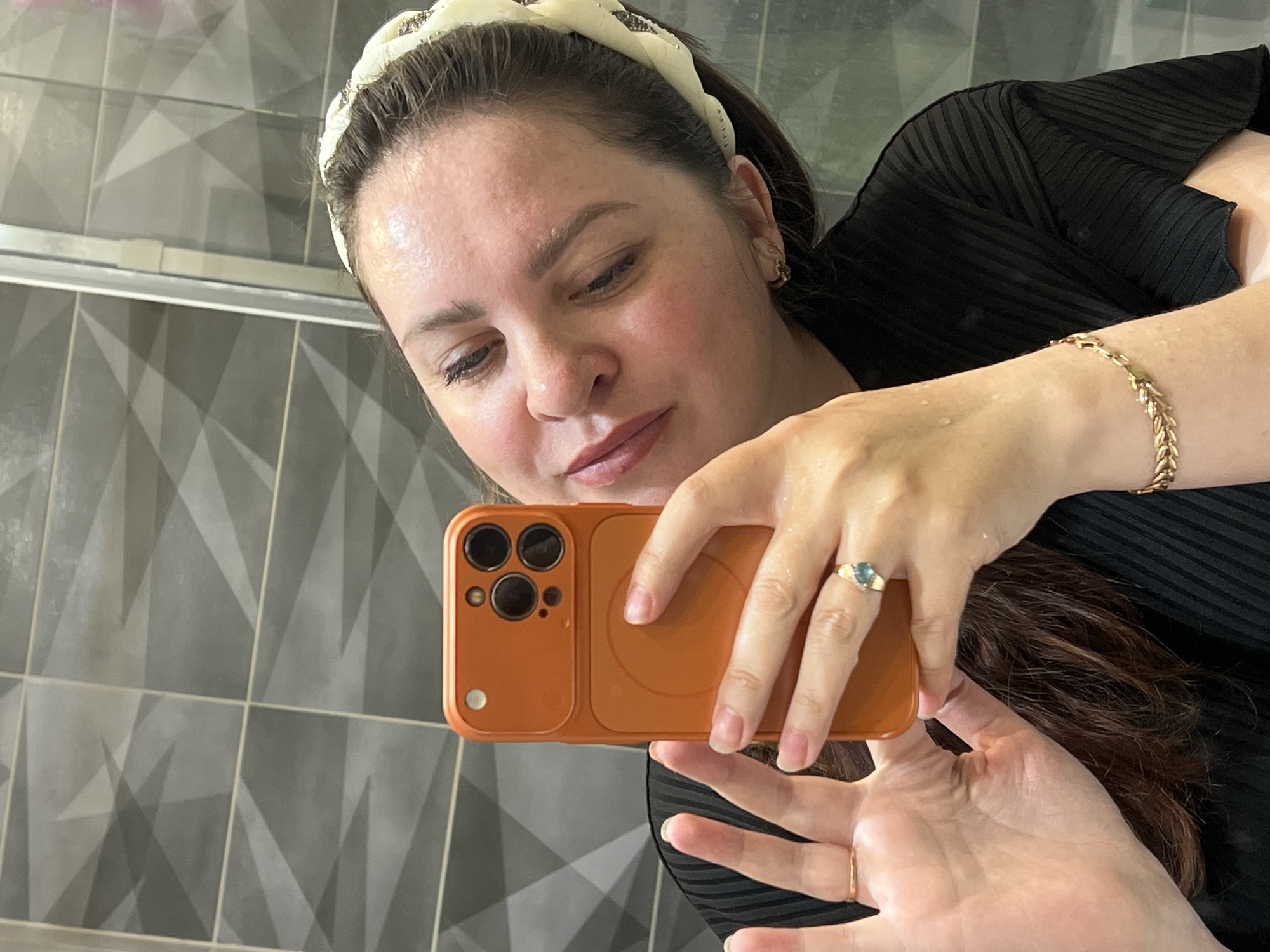An adult female is depicted in a mirror selfie taken indoors in the city of Noviy, Russia. The woman, who appears to be in her late 20s to early 40s, has light brown hair pulled back by a patterned light-colored headband. Her face and hands show small water droplets, suggesting recent exposure to water or high humidity. She wears a black, horizontally pleated top. On her left hand, a plain gold-toned ring is visible on her ring finger. On her right hand, a gold-toned ring with a prominent bluish-green gemstone is on her ring finger, and a gold-toned chain-link bracelet adorns her wrist. She is holding an orange-cased smartphone, with a circular magnetic accessory area and a three-lens camera module visible on the back, to capture the image. The background consists of grey wall tiles featuring a repeating geometric pattern of light and dark grey triangles. A vertical metallic frame, consistent with a shower enclosure or large mirror frame, is partially visible on the right side of the reflection.