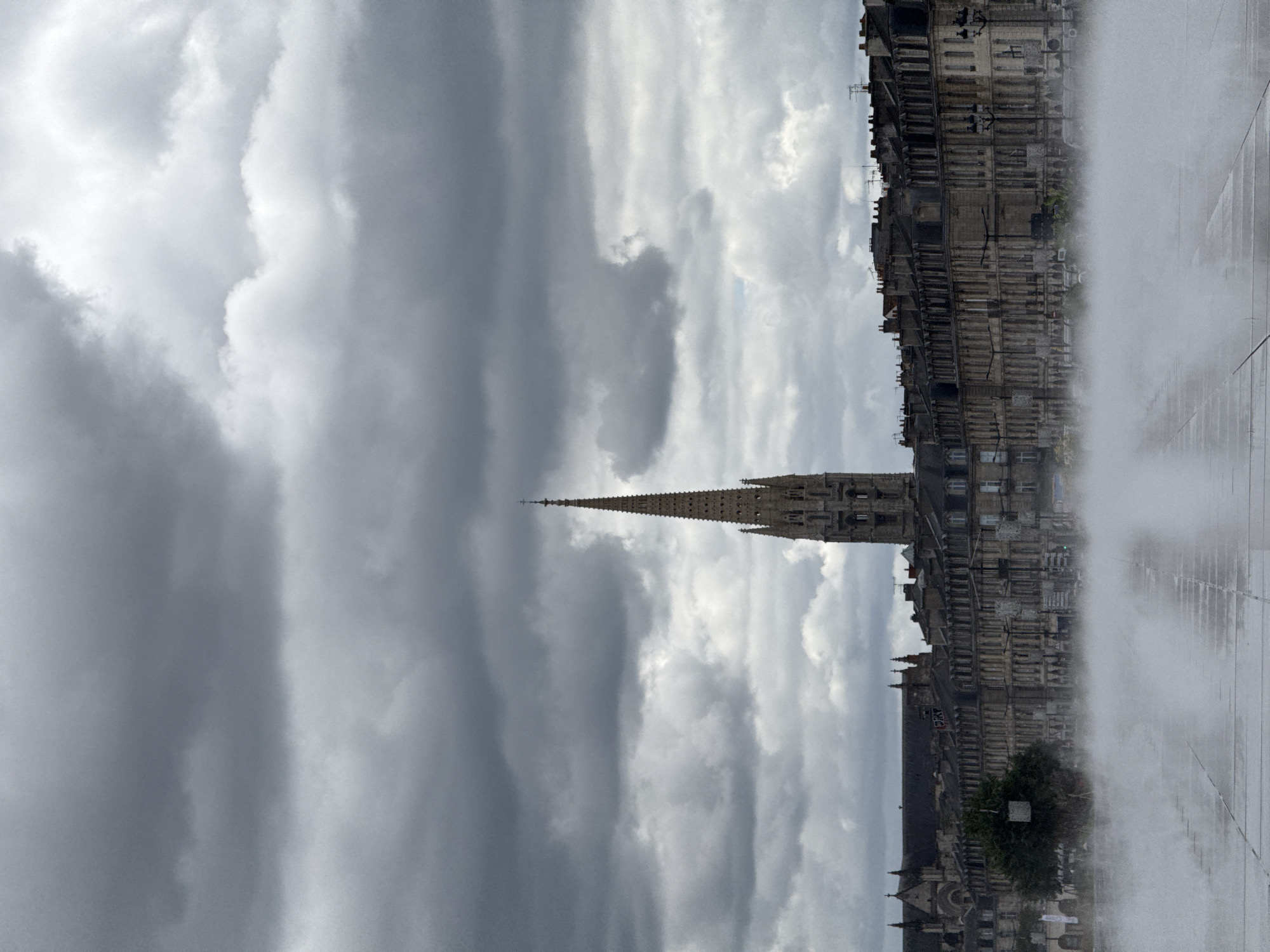 The image shows a wide-angle view of the Miroir d'eau in Bordeaux, France, on an overcast day. The foreground is dominated by a sheet of water with mist rising from its surface, reflecting the grey sky. Behind the water, a row of classical stone buildings stretches across the frame, their facades detailed with numerous windows and architectural elements. A prominent church spire, characteristic of Gothic architecture, rises above the buildings in the center of the image. The sky is filled with dark, dramatic clouds, suggesting impending rain or recent precipitation. The overall atmosphere is somber and grand, with the mist adding a sense of ethereal beauty to the urban landscape.