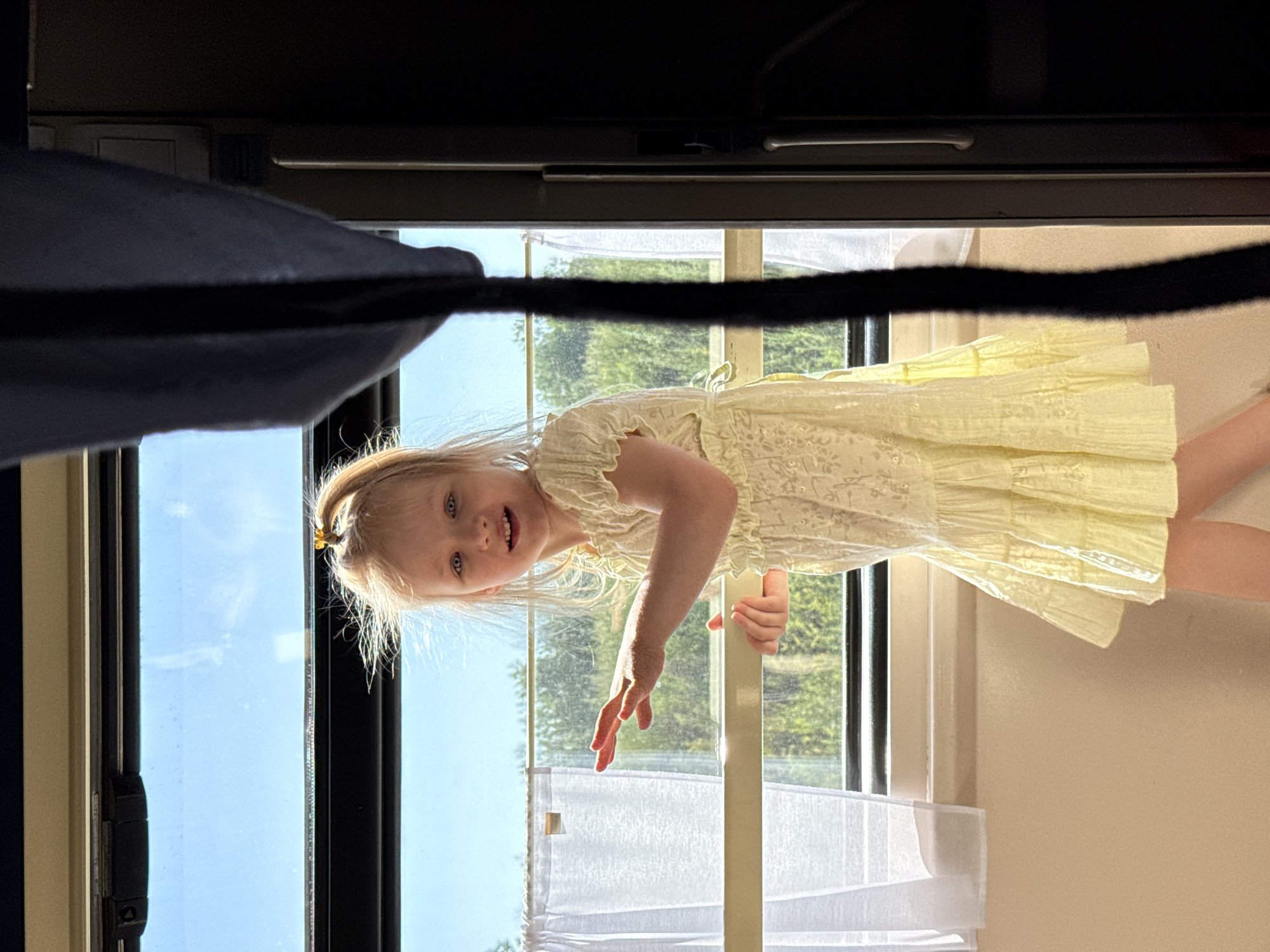 A young Caucasian girl with blonde hair tied in a ponytail, wearing a light yellow dress, is standing by a window with white curtains. She is looking at the camera with a slight smile and has her hands extended. Outside the window, trees are visible against a blue sky. The scene appears to be indoors, possibly in a train or bus, given the window structure and a dark curtain on the right side of the image. The location in Grushevskaya, Russia is not discernable from the image itself.