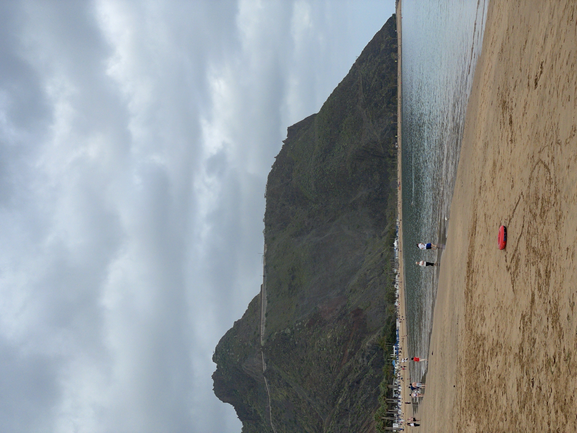 The image displays a serene coastal landscape, likely situated in Santa Cruz de Tenerife, Spain, under an overcast sky. The scene is dominated by a large, dark green, vegetated mountain that rises steeply on the left side of the frame, directly from the coastline. A faint, lighter-colored path can be seen winding its way up the mountain's face.
To the right of the mountain, a long, sandy beach stretches into the distance, meeting the calm, blue-green ocean water. The water shows gentle ripples and appears tranquil. Several people are visible along the shoreline; two individuals are seen wading in the shallow water near the center, and others are scattered further down the beach. In the background, along the base of the mountain, rows of beach chairs and blue umbrellas suggest a leisure or resort setting.
A distinctive red, U-shaped inflatable ring or buoy lies on the sand in the mid-right foreground. The sky above is entirely covered by soft, grey clouds, indicating an overcast day, which casts a diffused light over the scene. The overall activity appears to be one of relaxation and leisure by the sea. No visible text is identifiable within the image.