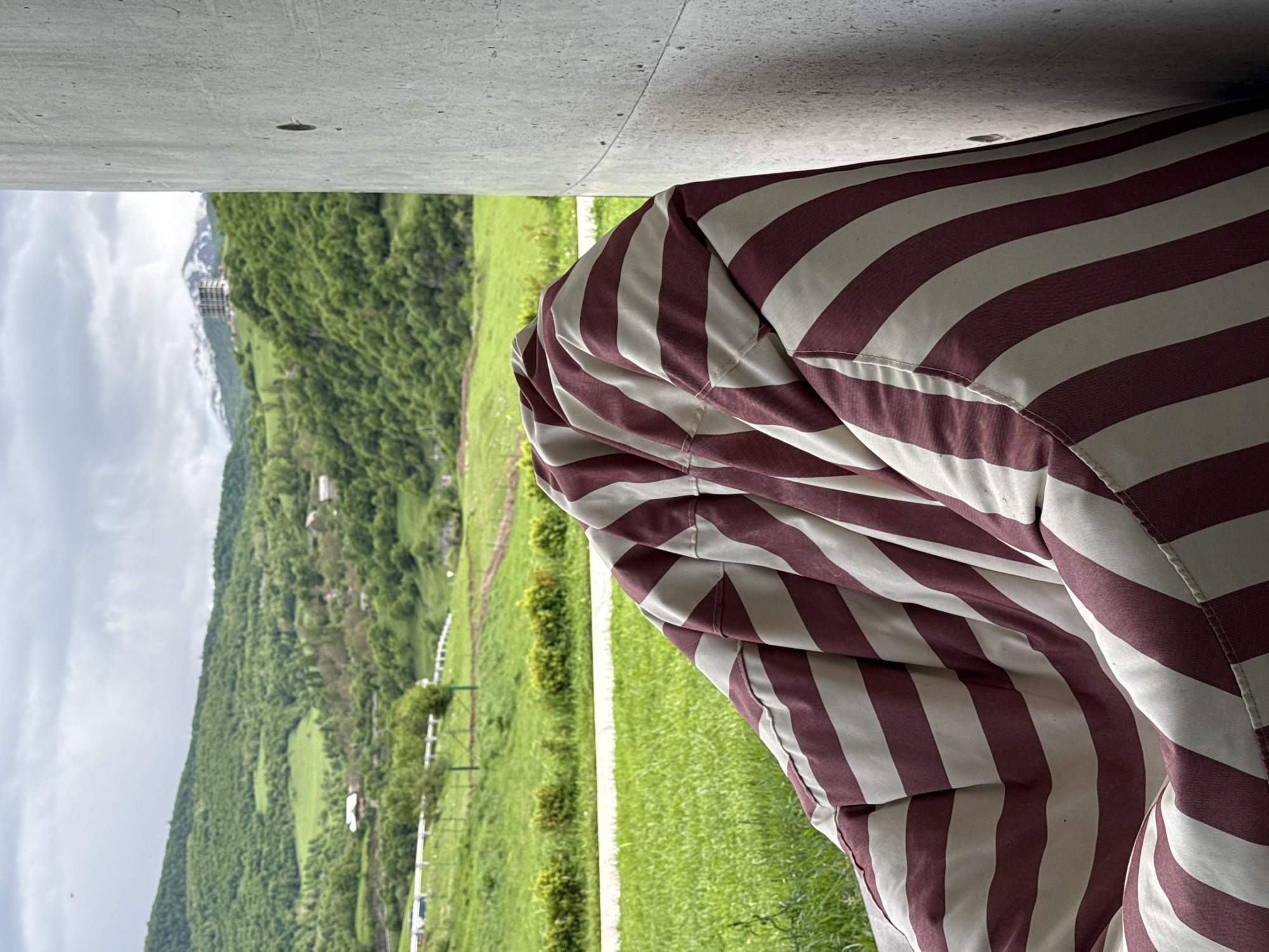 The image depicts a view from underneath a concrete structure, possibly a bridge or building overhang. The foreground is dominated by a striped beanbag chair in maroon and white. Beyond the concrete overhang and next to the beanbag chair is a landscape view featuring rolling green hills and mountains in the distance. There is a white fence visible in the mid-ground. The sky is overcast. The location is most likely Chkalov, Armenia.