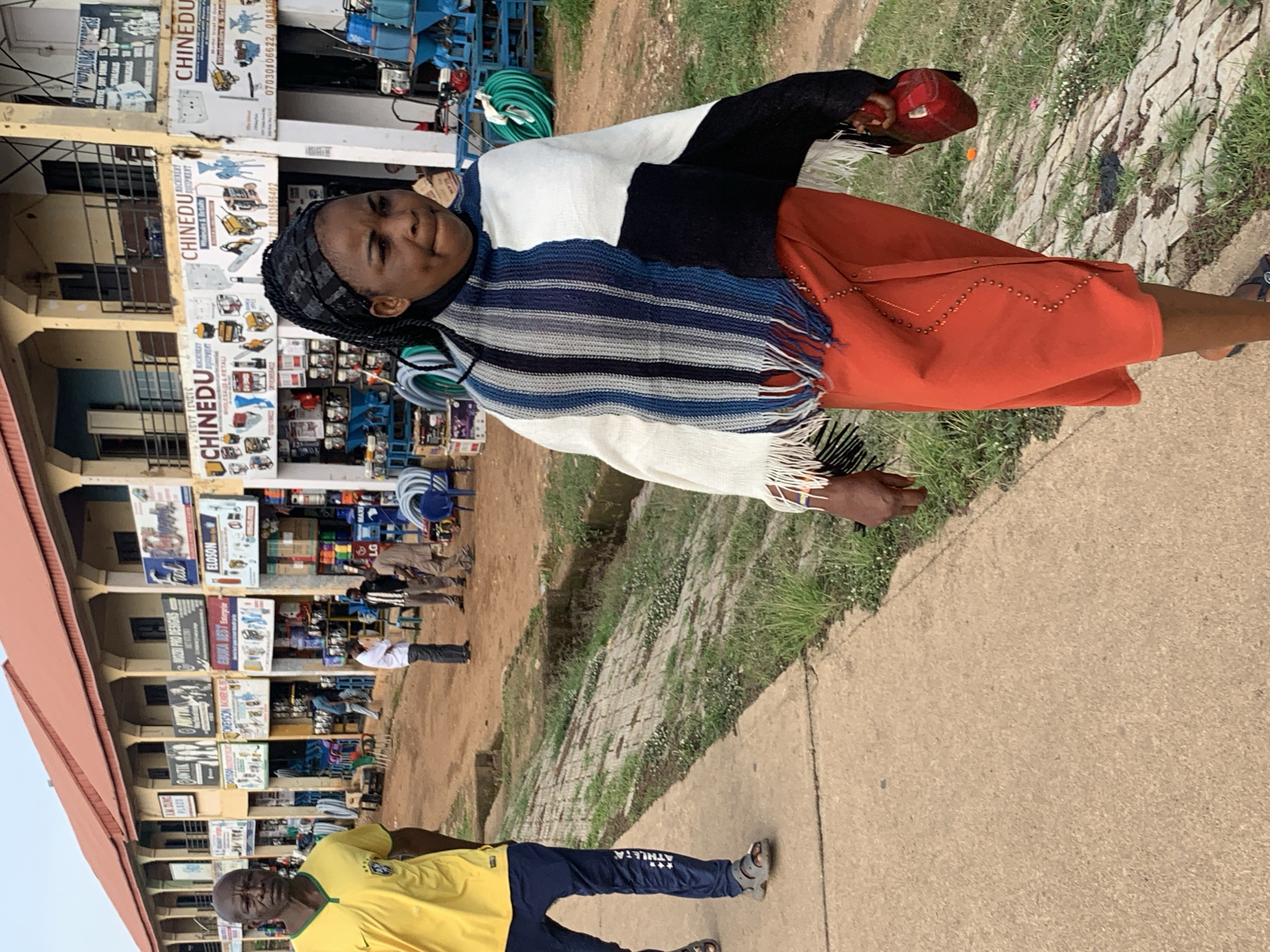 In the city of Gura Top, Nigeria, a woman with dark skin and braided hair walks along a paved sidewalk. She wears a black headscarf, a poncho-style garment with stripes of white, black, and varying shades of blue and gray, and a vibrant orange skirt adorned with silver studs. In her right hand, she carries a red bag. To her left, a man with dark skin and a closely cropped haircut walks in the opposite direction. He is wearing a bright yellow t-shirt with a dark collar and a dark blue tracksuit with "ATHLETA" printed on the leg. He is also wearing dark sandals. Behind the woman and man, a row of shops lines the street. The buildings have red roofs and are painted in shades of yellow and white. Signs for businesses such as "CHINEDU MACHINERY EQUIPMENT" are visible. Various items are displayed outside the shops, including machinery, hoses, and stacks of goods. Several other individuals are present near the shops, some appearing to be customers or shopkeepers. The ground is a mix of dirt and some paved areas, with patches of grass and a shallow ditch. The sky is overcast, suggesting a cloudy day.