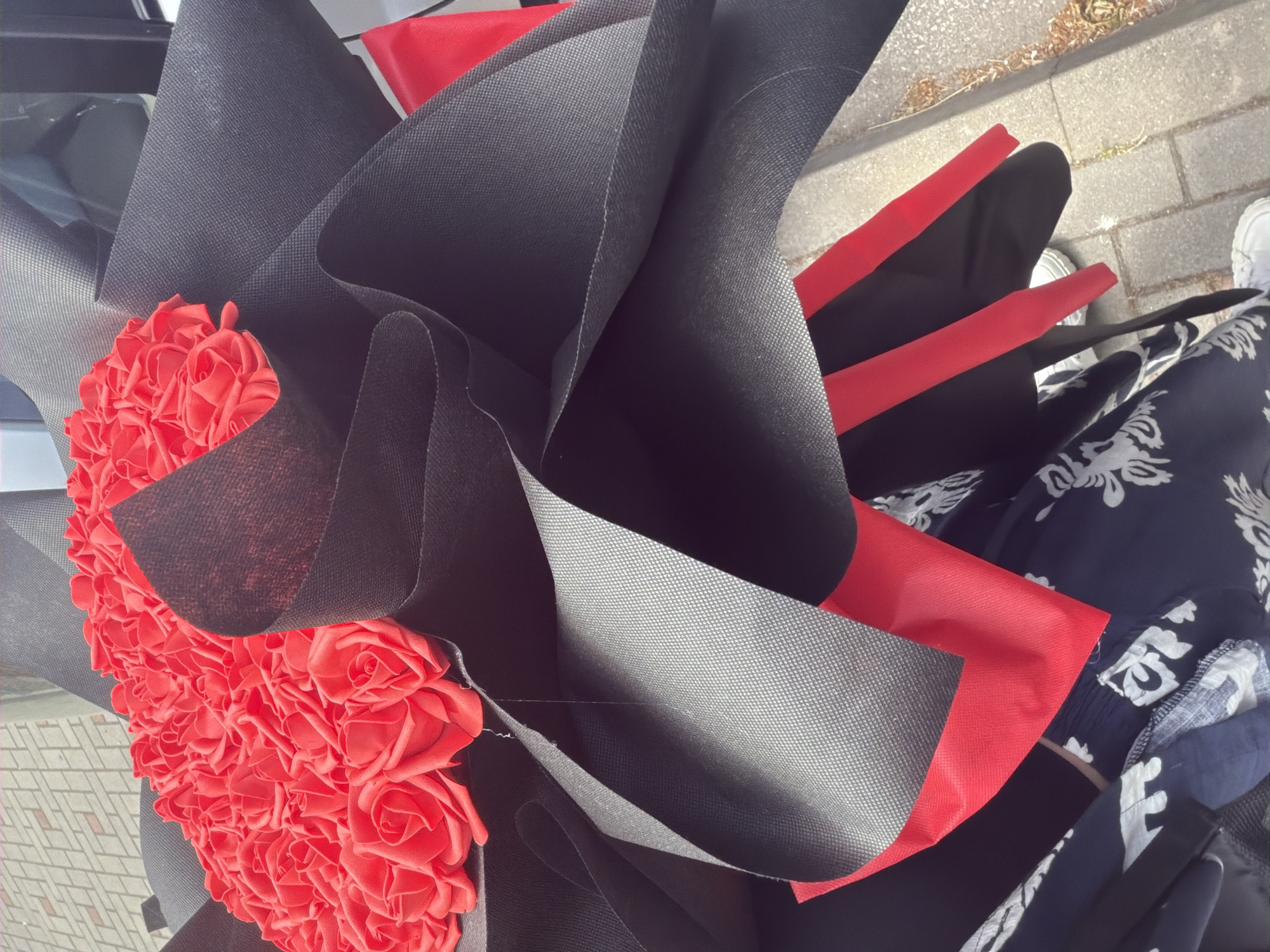 A bouquet of red roses wrapped in black and red paper is visible in the foreground. The bouquet is placed on what appears to be a person's lap, who is wearing dark blue pants with a white floral pattern. The lower part of the image shows a sidewalk made of grey paving stones and some dry grass. The scene is outdoors, likely in a city environment. No specific location in Pazardzhik, Bulgaria can be identified from the visual information provided.
