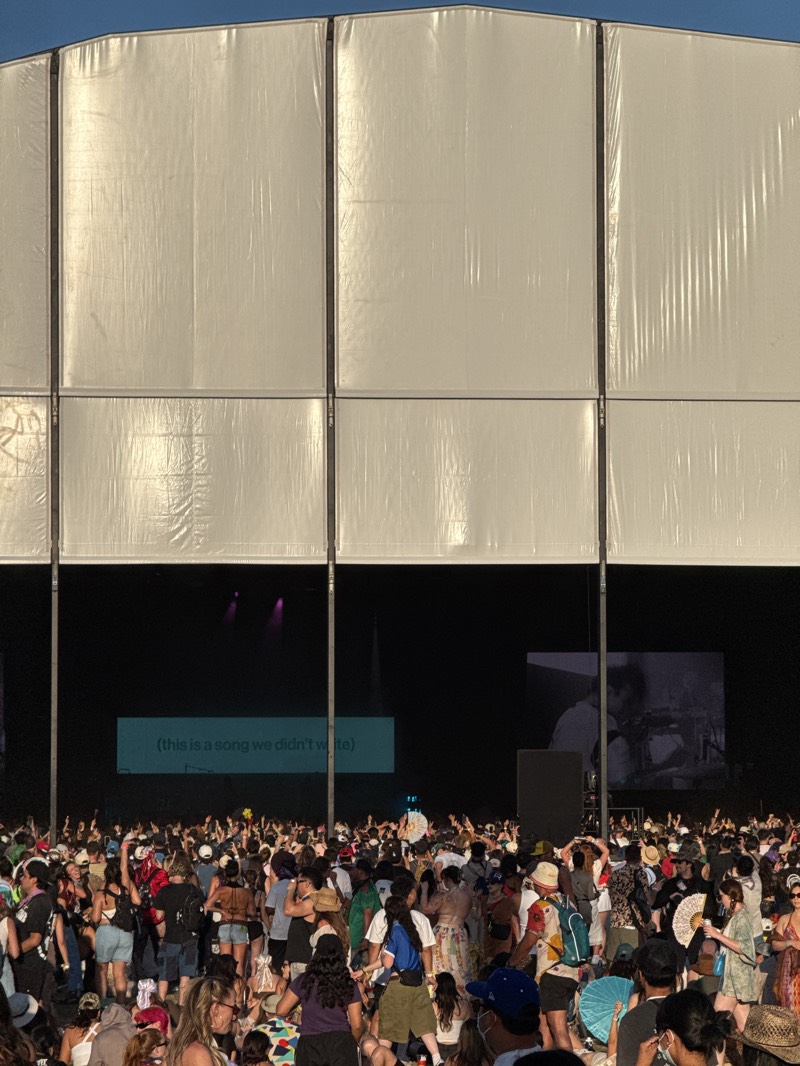 A large crowd attends an outdoor concert at what appears to be a music festival, likely in Indio, California, United States, given the environment and crowd appearance. A large white stage canopy covers the performance area. A screen displays the phrase "(this is a song we didn't write)". The crowd is densely packed with people of various ages and ethnicities, many holding their hands in the air. Some individuals wear hats, and others use fans. The overall environment suggests a sunny outdoor event with music and entertainment.