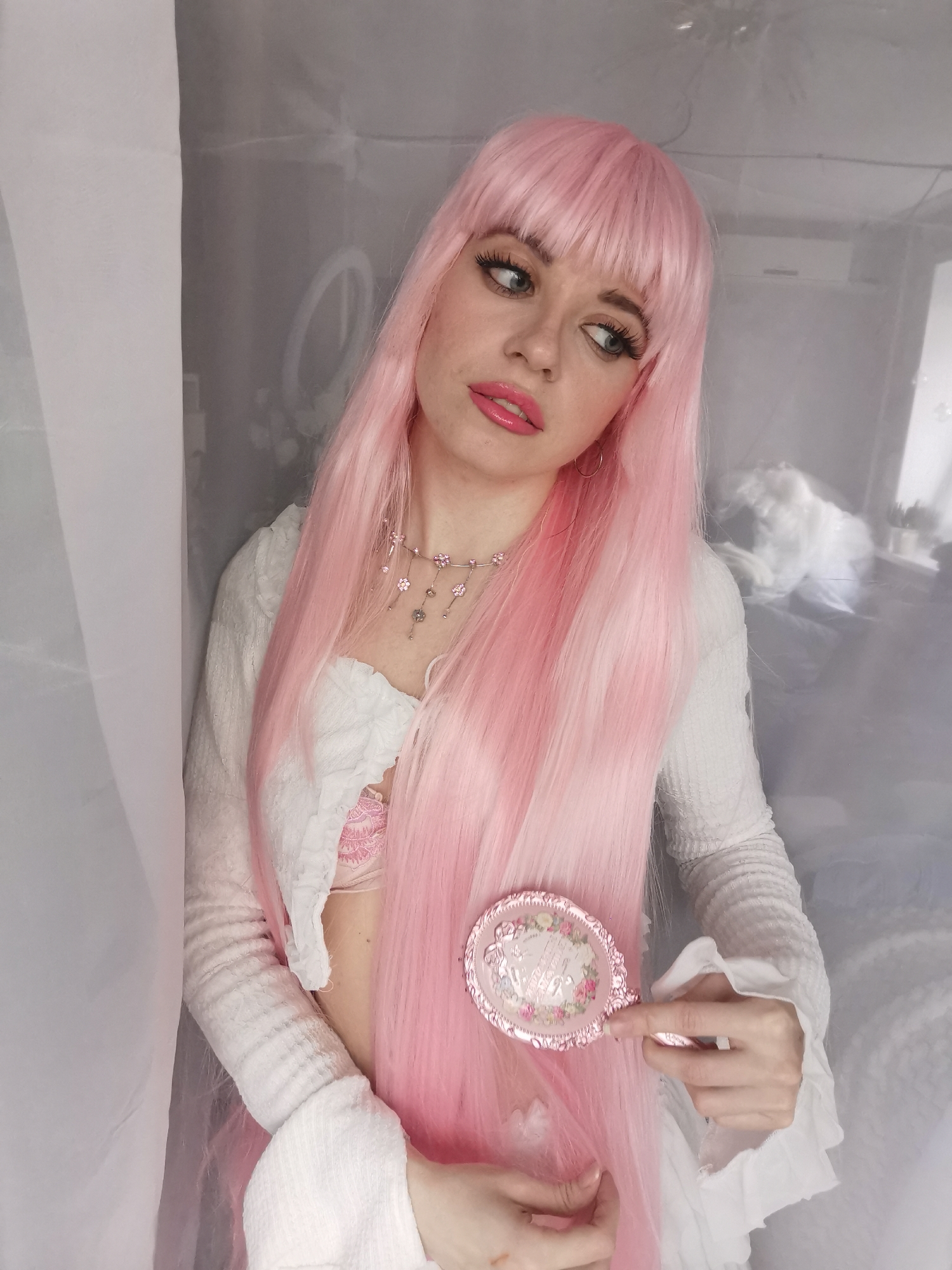 A young individual with fair skin and blue-green eyes is featured in a medium close-up, looking upwards and to the right with a pensive expression. They wear a long, bright pink wig with bangs, styled to fall over their shoulders. Their lips are painted with glossy pink lipstick, and their eyes are enhanced with prominent eyelashes.
The subject is dressed in a white, long-sleeved, cropped top made of a textured fabric, possibly ribbed or lace, which exposes their midriff. A pink lace bra or bralette is visible beneath the top. A delicate silver-toned necklace with small pink floral and clear dangling charms adorns their neck, and a gold hoop earring is visible on their left ear.
In their right hand, they hold a small, ornate, pink handheld mirror decorated with a floral design, positioned near their pink hair.
The setting appears to be indoors. On the left side of the frame, a vertical panel of white, sheer, translucent fabric, likely a curtain or screen, partially obscures the background. The background itself is softly blurred but reveals elements of a room through a transparent or reflective surface, possibly a sheer net or glass. Visible details include a ring light, an air conditioning unit, and indistinct forms suggesting furniture or bedding.
The lighting is soft, creating a gentle and ethereal atmosphere around the subject. The overall impression is that of a posed portrait, with the subject's gaze conveying a thoughtful or contemplative mood. No discernible text is visible in the image.