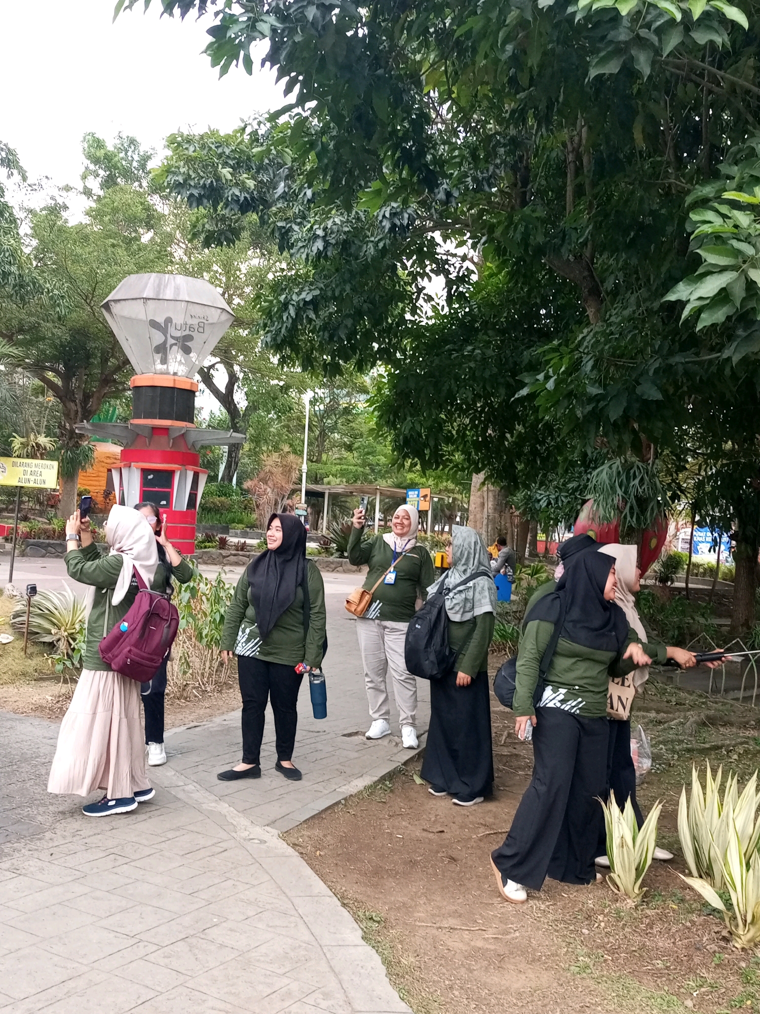 The image captures a group of women in casual outdoor attire, primarily consisting of olive green tops and headscarves, standing on a paved walkway bordered by soil and vegetation. The scene appears to be set in an urban park or recreational area. In the background, a distinctive, modern-looking structure with a diamond-shaped top is visible, partially obscured by trees. A sign in Indonesian, "DILARANG MEROKOK DI AREA ALUN-ALUN" which translates to "NO SMOKING IN THE TOWN SQUARE AREA", suggests the location is a town square in Batu, Indonesia. Several women are holding smartphones, with at least two appearing to be taking photos or videos. One woman is using a selfie stick. The overall atmosphere is relaxed and informal, with the women engaged in conversation and activities. The environment is characterized by lush green trees and undergrowth, indicating a temperate climate.