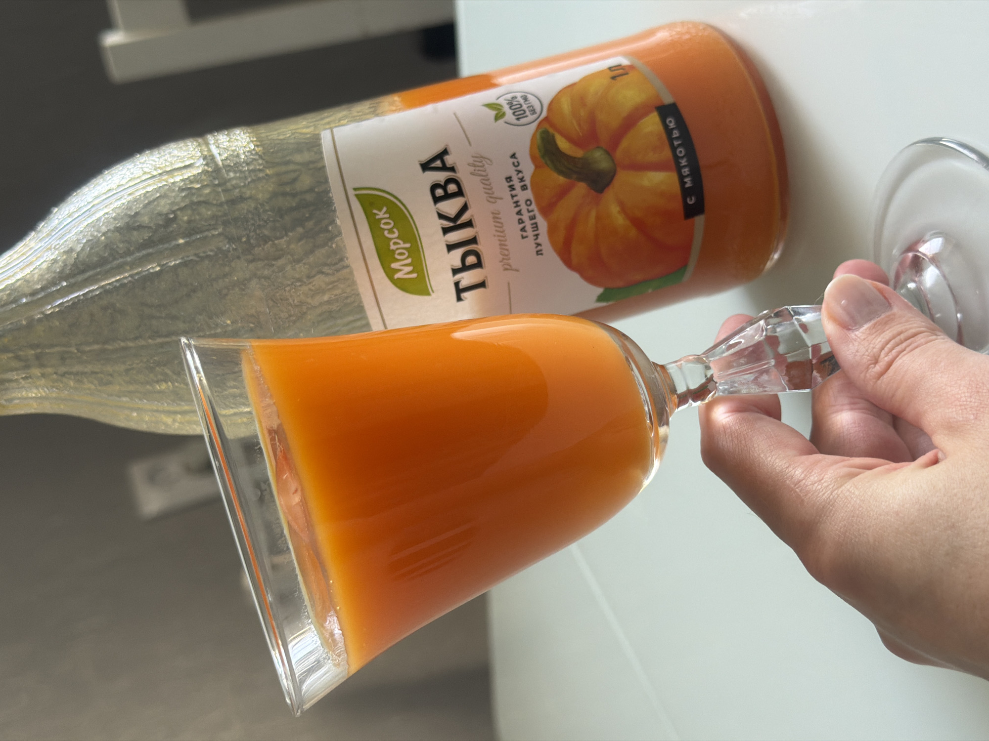 The image shows a bottle of pumpkin juice (Тыква) from the Russian brand "Morsok" lying on its side, next to a filled glass being held by a person's hand. The bottle label features a pumpkin and text in Russian, indicating "premium quality" and "guarantee of the best taste." The text also says "with pulp". The glass is a decorative crystal glass, filled with the orange juice. The background appears to be a white surface. While the image contains Russian text, there is no direct information or visual evidence to indicate this image was taken in Rostov-on-Don, Russia.