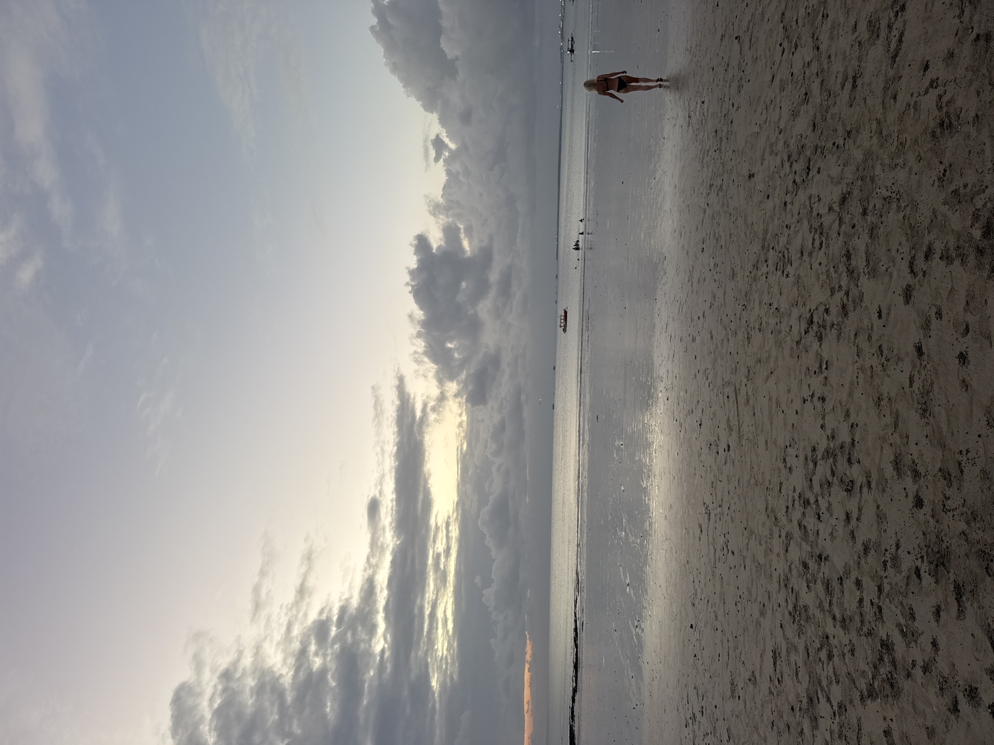 A wide shot captures a serene beach scene in Kuta Selatan, Indonesia, at dusk. The sky above is a canvas of soft grays and whites, transitioning to a pale yellow glow near the horizon where the sun has set. Dark, textured clouds drift across the sky, with lighter, wispy clouds interspersed. The ocean stretches out, appearing calm and reflective, with a subtle sheen mirroring the sky.
In the foreground, a sandy beach is wet from the receding tide, dotted with small pebbles and shell fragments. Footprints mark the damp sand, indicating recent activity. A lone woman, wearing a black bikini, walks away from the camera towards the water, her blond hair visible.
In the middle ground, several small boats are scattered across the water. Some appear to be anchored, while others are in motion. A group of people are visible near one of the boats, some standing in the shallow water. Further out, a few more boats are visible as distant silhouettes. The overall atmosphere is peaceful and tranquil, with the approaching evening casting a soft, diffused light over the landscape.