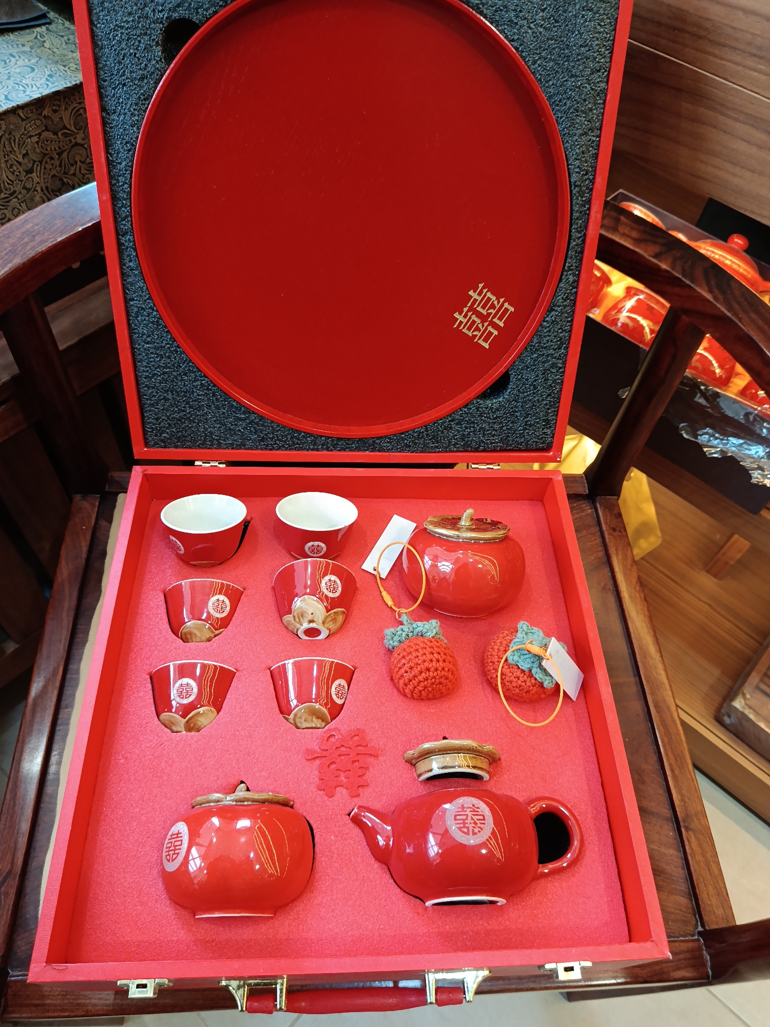This image shows a red Chinese tea set in a matching red box, likely for a traditional tea ceremony. The set includes several small teacups, two teapots, a round serving tray, and two decorative red strawberry-shaped ornaments. The tray and the teapots are marked with a Chinese symbol, possibly for "double happiness". The set is placed in a red box with a foam insert that is resting on a chair. Based solely on visual content, there is no indication of its location, and it is not possible to establish that this image was taken in Moscow, Russia.