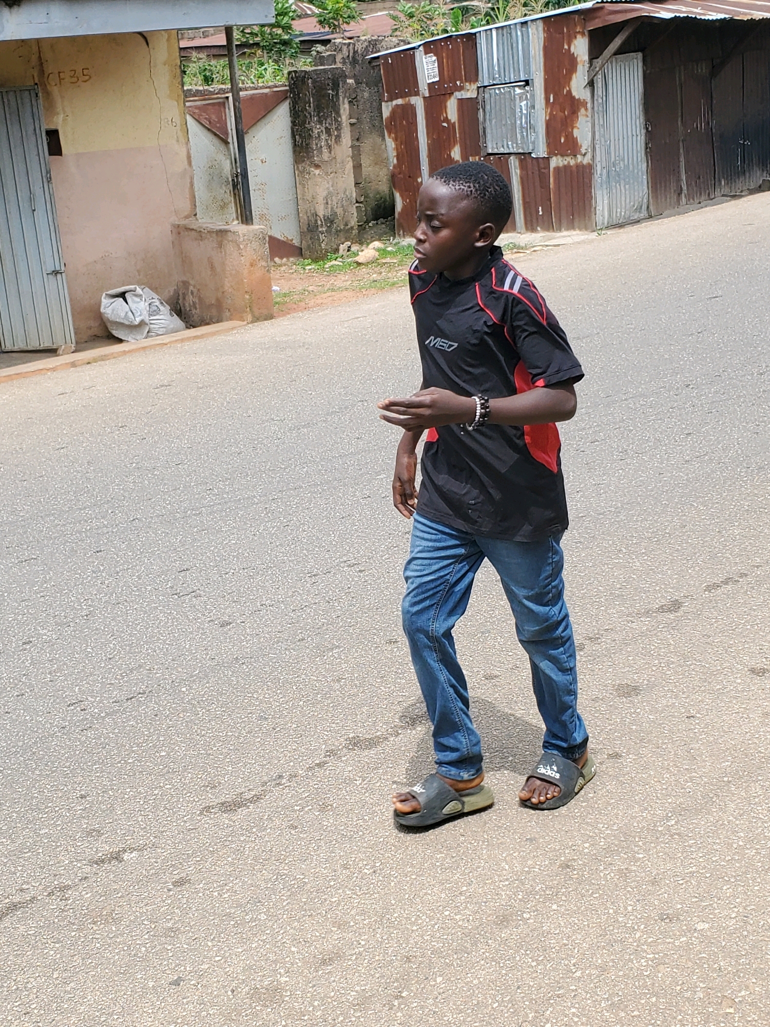This media file depicts a young Black boy walking on a paved road in what appears to be a semi-urban or rural setting in Jos, Nigeria. The boy is wearing a black t-shirt with red accents and the text "MED" visible on the front, blue jeans, and dark slide sandals. He has bracelets on his left wrist.
The setting includes a weathered building with a faded "CF35" marking on its wall and a large, gray plastic bag resting near its entrance. Adjacent to this building, there is a corrugated metal structure, possibly a shop or dwelling, with a door and a window. In the background, there are glimpses of vegetation and more corrugated metal structures, suggesting a residential or commercial area. The road surface is asphalt with visible markings and some debris.
The lighting suggests it is daytime, likely sunny, given the shadows. The boy's gaze is directed to his left, and his posture indicates he is in motion. The overall impression is of a candid shot capturing a moment in the daily life of a child in this specific location. The visible text "CF35" and "MED" are present on the building and the boy's shirt, respectively.