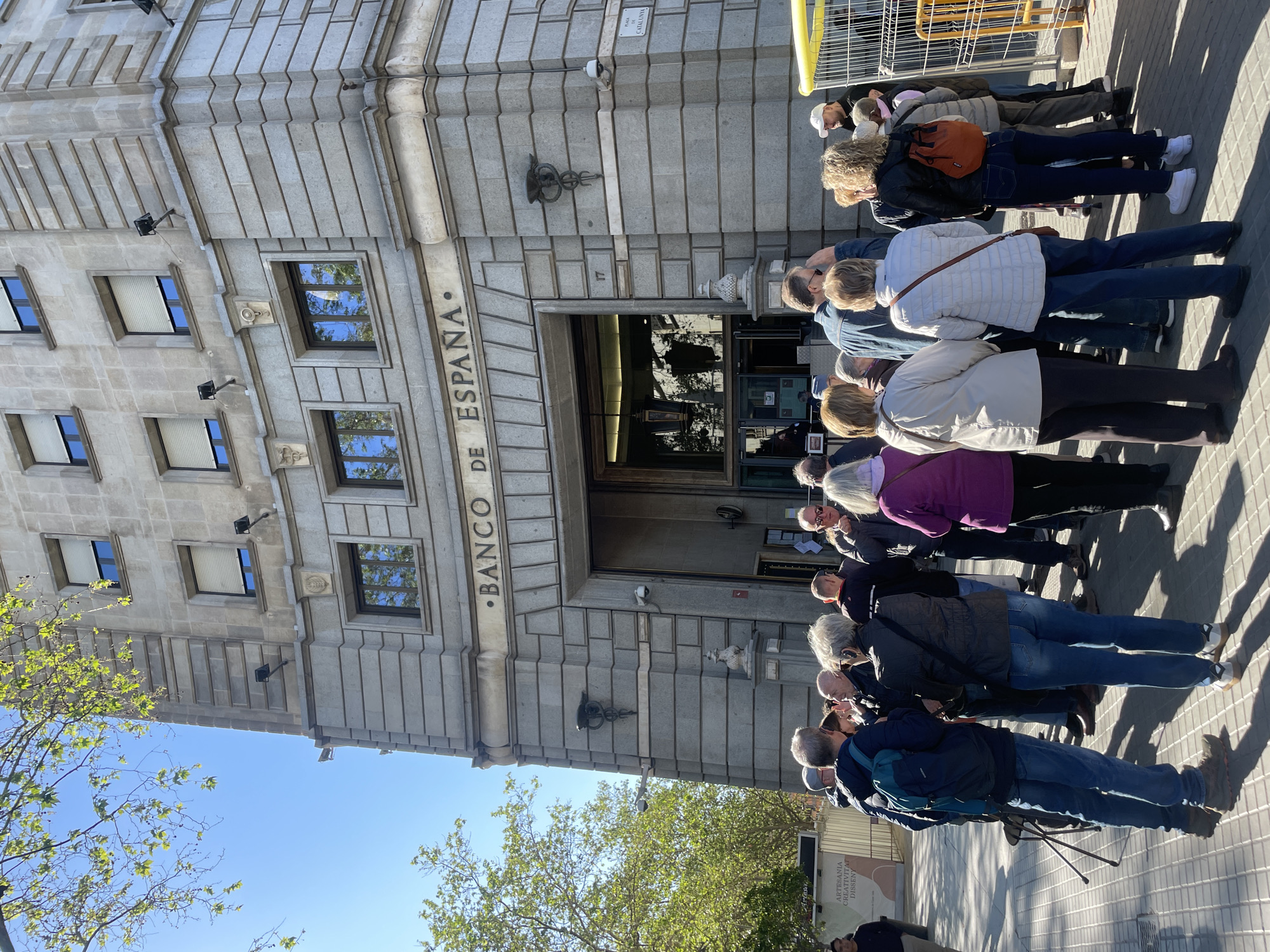 A horizontal image depicts a group of people standing in line in front of a building labeled "BANCO DE ESPAÑA." The building, likely a bank or financial institution, is made of light grey stone or concrete and has multiple windows. A sign with the number "17" is visible near the entrance. The people are dressed in casual clothing, suggesting a daytime scene. The pavement is visible in the foreground. The location is Madrid, Spain.