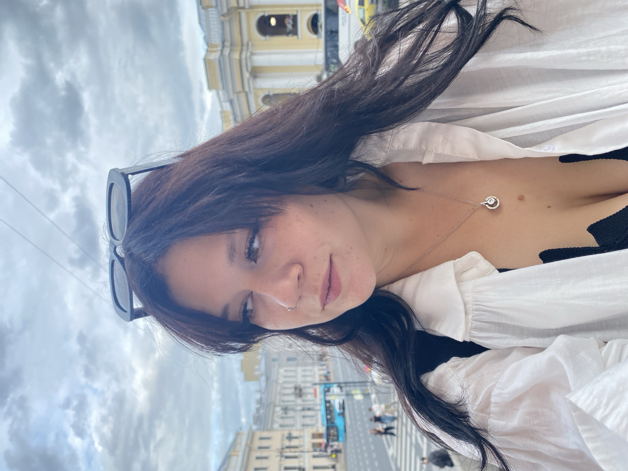 A young woman with dark hair and sunglasses perched on her head takes a selfie outdoors. She is wearing a white, voluminous-sleeved shirt over a black top, and a silver necklace with a pendant. Her nose is pierced with a hoop ring. The background shows a city street scene, with buildings, a blue bus, and people walking on a crosswalk. The architecture suggests it is the city of Saint Petersburg, Russia, possibly near a prominent landmark like the Winter Palace or Admiralty Building, given the classical style of the yellow and grey buildings. The sky is overcast with clouds.