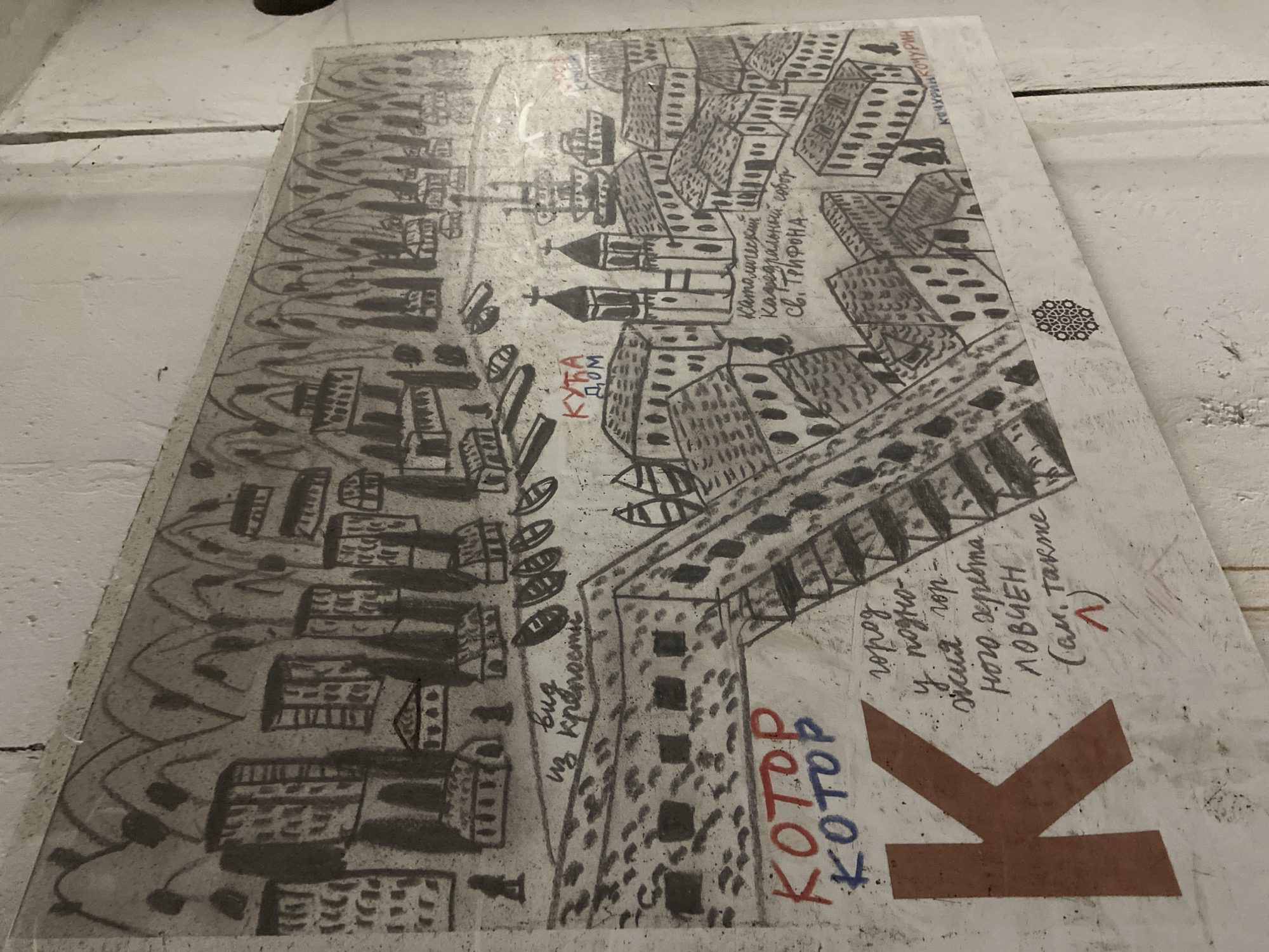 The image shows a hand-drawn, possibly charcoal or pencil, artistic rendering of the city of Kotor, Montenegro. The drawing depicts the city layout, including buildings and the bay, with some written annotations in what appears to be Russian or a related Slavic language.
The lower right corner features a large, brown "K" shape, likely for Kotor. Adjacent to it are the words "KOTOP" in both red and blue. Further above, hand-written text describes Kotor as a town at the foot of the Lovćen mountain range.
The drawing includes representations of buildings, including the Catholic Cathedral of Saint Tryphon, and features a depiction of the Bay of Kotor. The handwriting indicates "A view of the fortress".