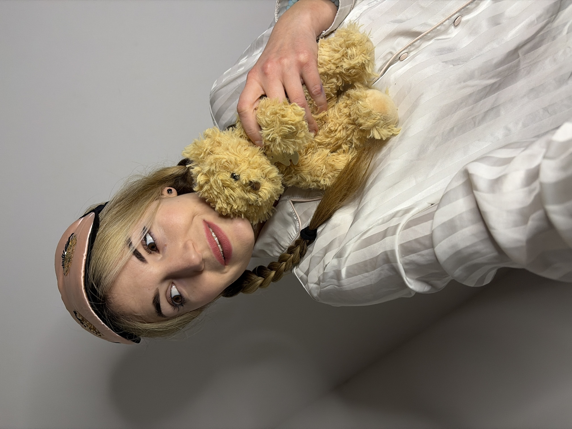 This image captures a fair-skinned female individual indoors in Budva, Montenegro. She possesses blonde hair styled into two visible braids, a pink sleep mask featuring gold sequin eyelashes positioned on her forehead, and a black ear stud in her left ear. She wears a light-colored, vertically striped satin pajama top with pink piping along the button placket. Her lips are colored with pink lipstick, and she exhibits a slight smile while gazing upward and to the left of the frame. She holds a small, light-brown, fluffy teddy bear against her chest with her left hand, which has a small metallic tag attached to its neck. The background consists of a plain, light grey wall, illuminated by soft, even lighting.