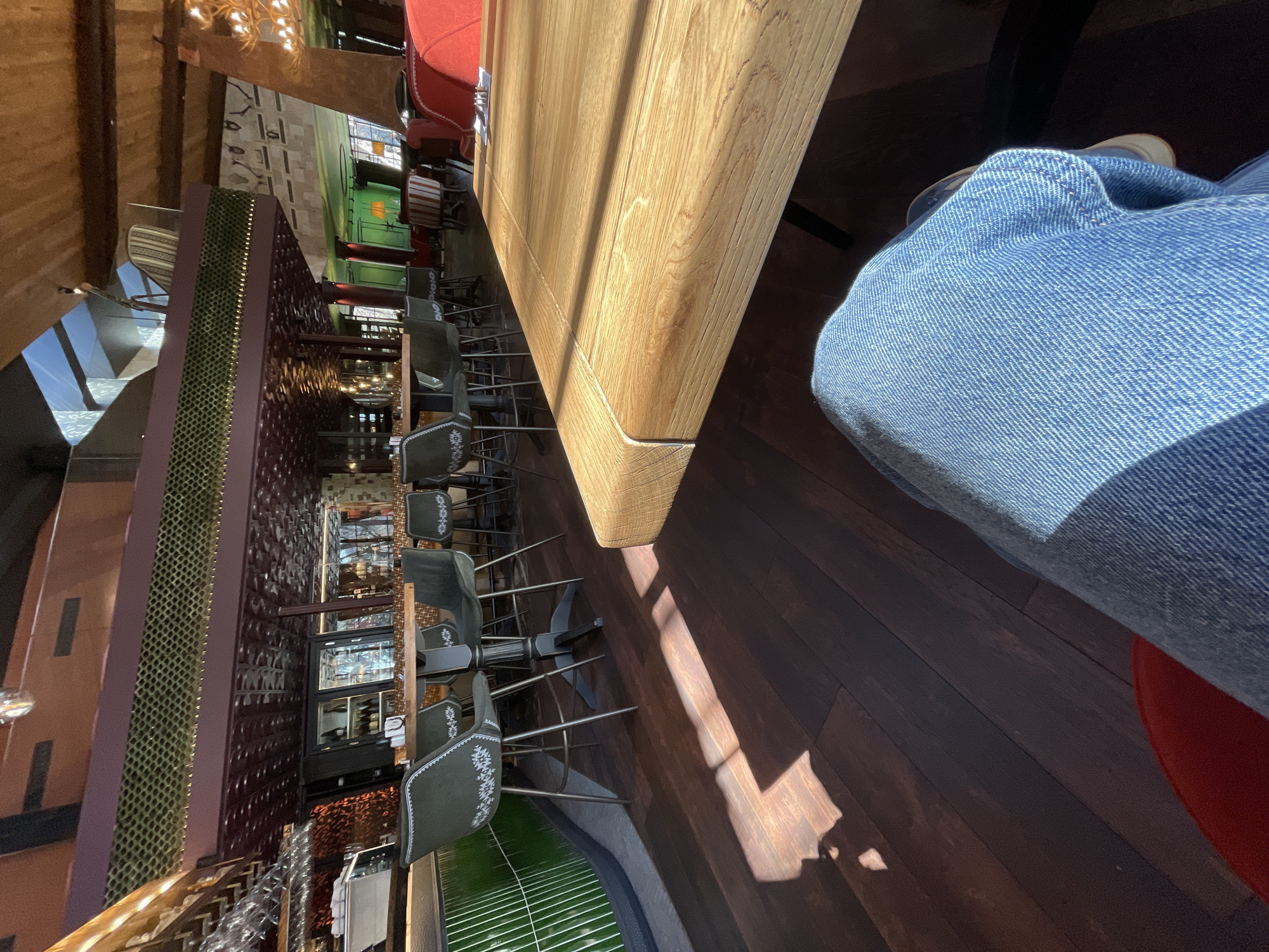 The image shows an interior space in Sofia, Bulgaria, likely a bar or restaurant. A long wooden table dominates the right side of the frame, angled slightly away from the viewer. Stools with patterned upholstery are positioned along one side of the table, and a person wearing blue jeans is partially visible at the right edge. A red chair back is visible. A dark, wood-planked floor extends beneath the table and stools. On the left is a mirrored wall, reflecting bottles and glassware, with a brown lattice-like structure dividing the mirrored area. A green, curved structure with vertical slats is located at the far left. The overall lighting suggests daylight entering the space.