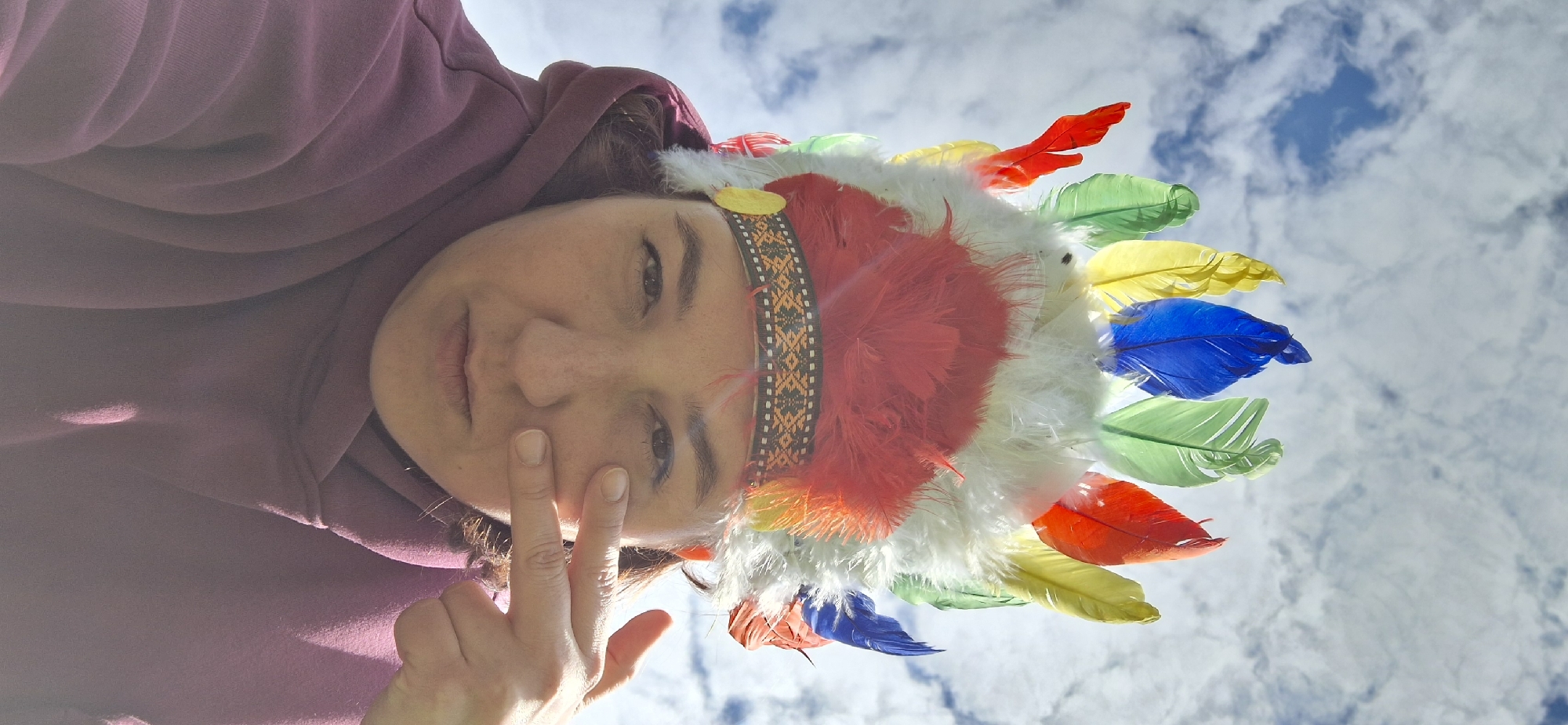 A woman wearing a feathered headdress and a maroon hoodie is posing in front of a blue sky with clouds. She is looking at the camera and holding two fingers to her face. The location is unspecified, not necessarily Прикамский, Russia.