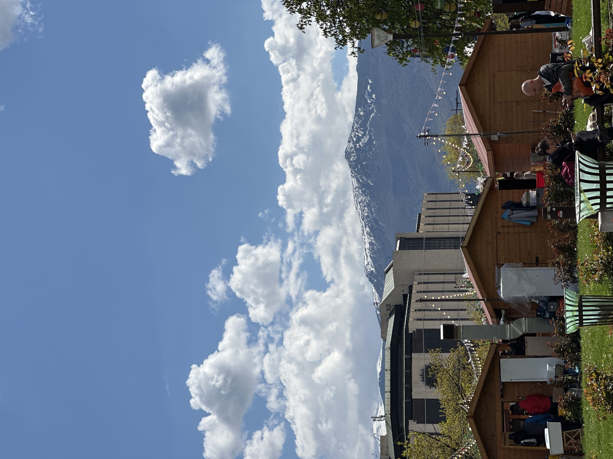 An outdoor scene captures a vibrant market or festival area set against a dramatic mountainous backdrop. The image is taken on a bright, sunny day, featuring a clear blue sky dotted with fluffy white cumulus clouds.
In the foreground and midground, several rustic wooden stalls or cabins with brown roofs are visible, suggesting a market setup. Green grass and bushes with reddish foliage frame these structures. People are scattered throughout the scene; a bald man in a striped shirt stands near one stall, looking towards the right, while others are seated on green-striped benches or mingling around the stalls. Festive string lights and small white triangular bunting flags are strung between trees and posts, enhancing the celebratory atmosphere.
Behind the stalls and trees, modern, multi-story buildings with facades of glass and concrete rise. Dominating the background is a majestic mountain range, partially covered with snow, which adds a sense of grandeur to the urban and market setting. The presence of both urban architecture and natural landscapes suggests an active area in a city with close proximity to mountains. The overall impression is one of a pleasant outdoor gathering or event. No discernible text is clearly visible on any signs or objects. Given the context, this scene is likely in Sofia, Bulgaria.