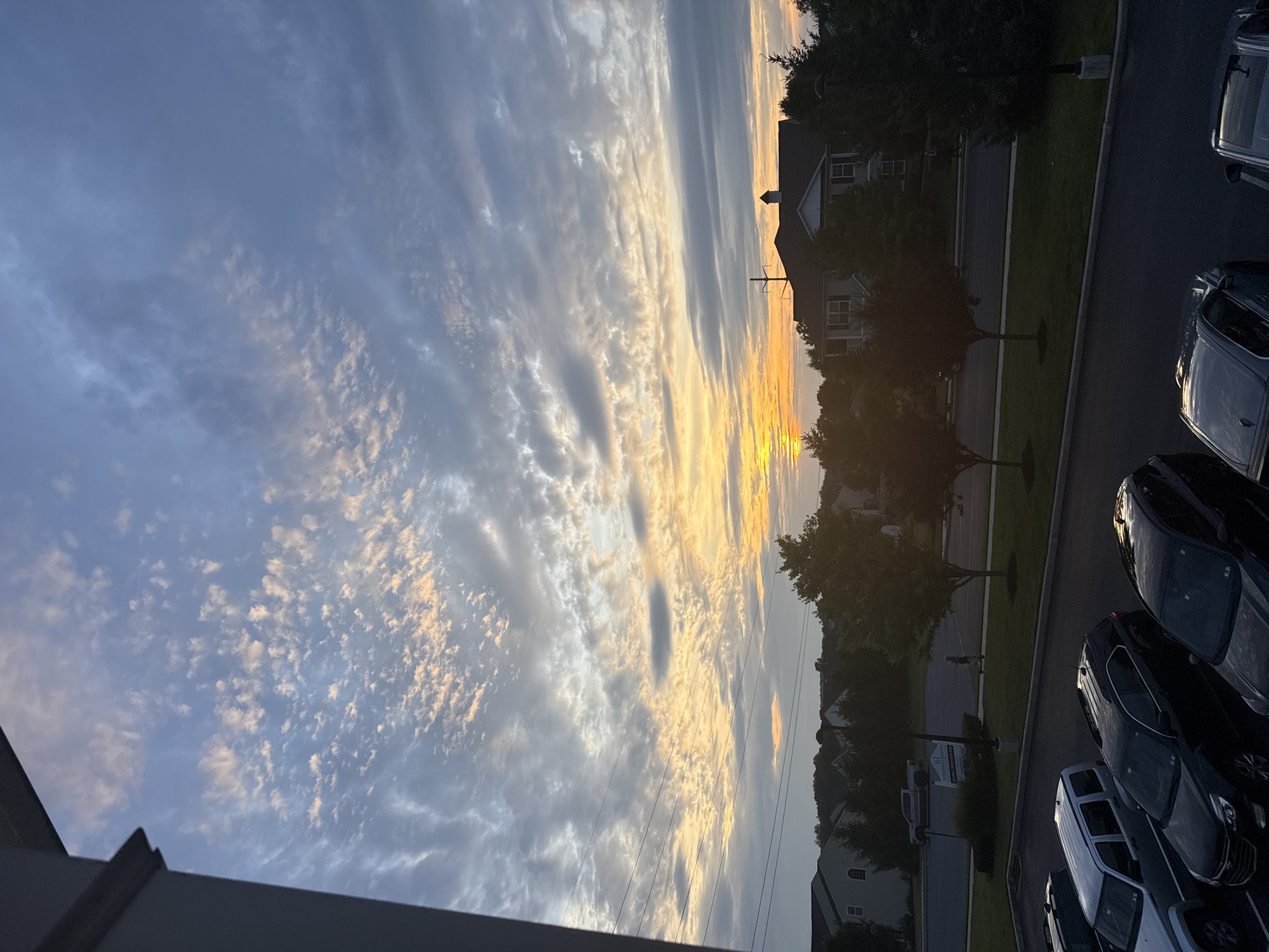 The image captures a scenic view of a sunset over Allen Township, United States of America. The sky is adorned with a mix of cirrus and cumulus clouds, painted in hues of orange, yellow, and blue, suggesting the sun is near the horizon. Silhouetted against the vibrant sky are several residential buildings and lush green trees. In the foreground, a parking lot is visible, occupied by several parked cars, including silver, black, and gray SUVs. A person is seen walking on a sidewalk in the middle ground, appearing to be enjoying the evening. The overall atmosphere is serene and peaceful, with the fading daylight casting a warm glow on the landscape.