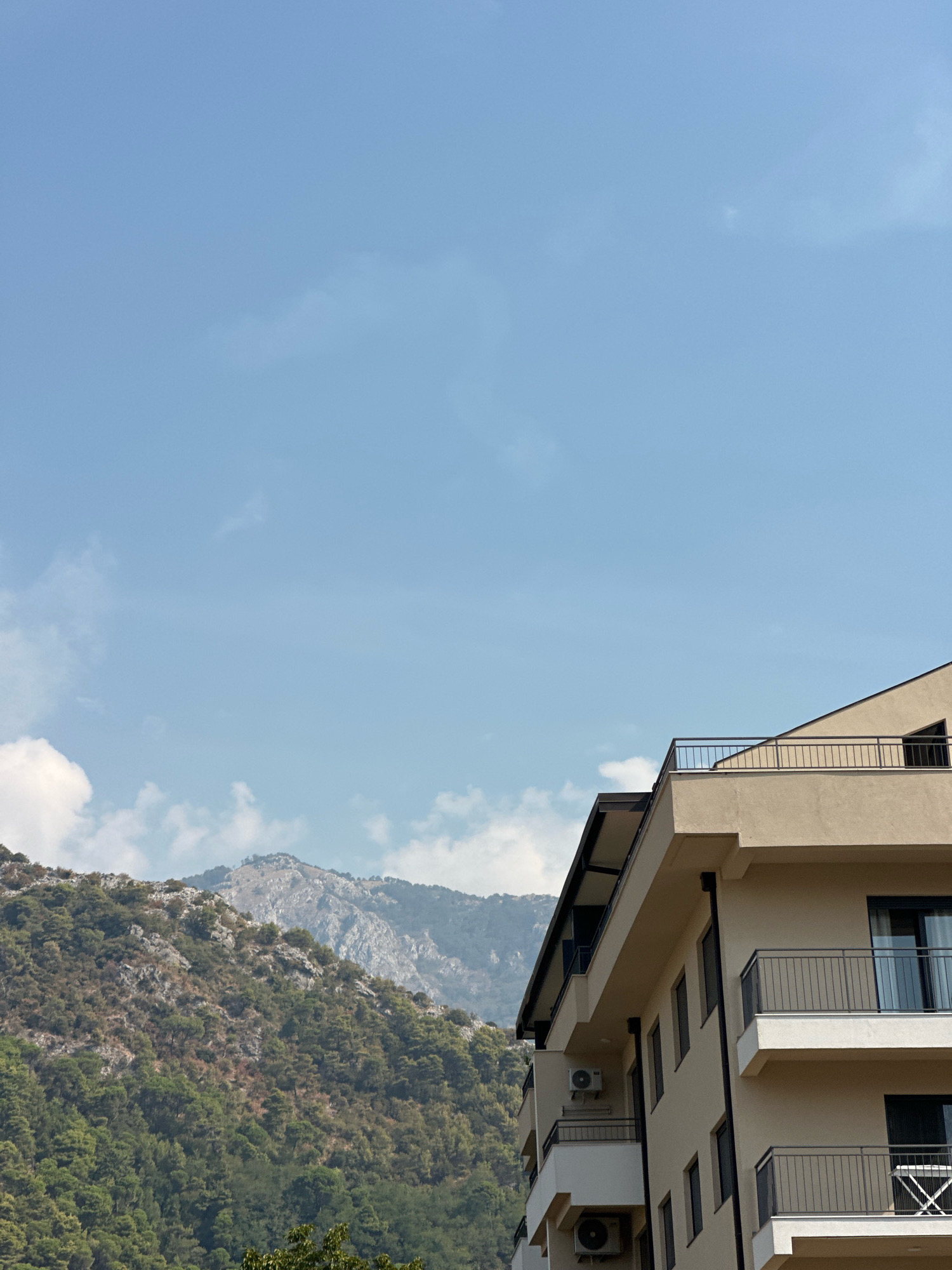This image, taken in Budva, Montenegro, features a modern, beige-colored apartment building in the foreground, partially obscuring a lush, mountainous landscape. The building has multiple balconies with black railings and dark-framed windows. An outdoor air conditioning unit is visible on one of the building's levels. The mountains in the background are covered with dense green vegetation and rocky outcrops, with a hazy, lighter-colored peak visible further in the distance. The sky above is a clear, pale blue with a few scattered white clouds.