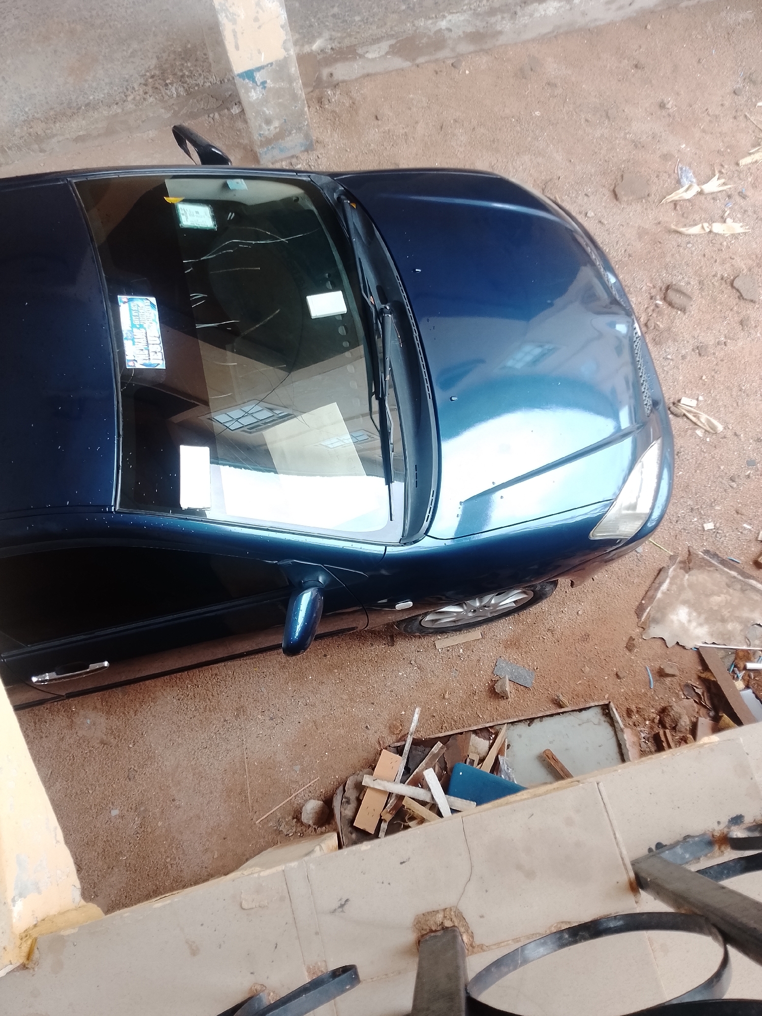 The image shows a dark blue car parked on a dirt and gravel surface. The car is viewed from a high angle, looking down onto the roof and front of the vehicle. The car appears to be a sedan or coupe with a sunroof. The windshield is visible, and there are some papers or stickers on it. A side mirror is also visible on the driver's side. The surrounding area is rough and appears to be an outdoor space, possibly a parking lot or an informal yard. There are some scattered pieces of wood and debris on the ground. The lighting suggests it is daytime, and the weather is likely clear, as there are reflections on the car's surface. No people are visible in the image. One sticker on the windshield has text that appears to be in a foreign language, possibly related to registration or inspection. Another sticker also appears to have some writing.