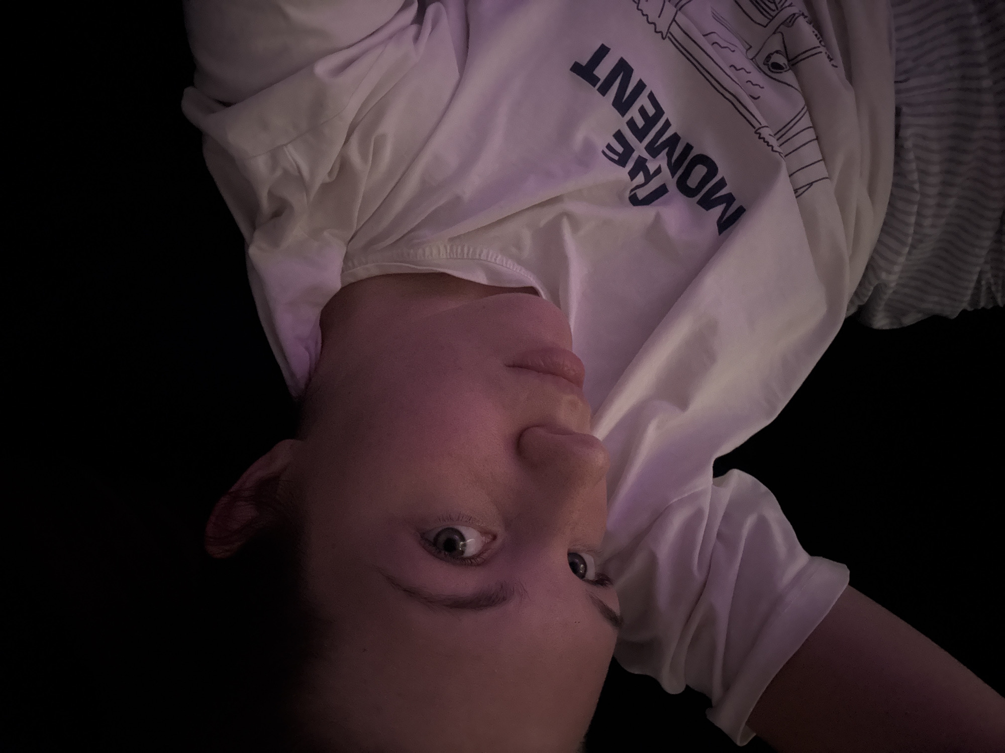 A low-angle, close-up shot captures a person's face and upper body against a dark background. The person, appearing to be female with fair skin and light-colored eyes, is looking upwards towards the camera, with their head tilted to the side. Their hair is dark brown and pulled back, with some strands visible on the forehead. A soft, purple light illuminates the left side of their face, casting shadows on the right. They are wearing a white t-shirt with a graphic design and text that reads "THE MOMENT" in dark blue lettering. The t-shirt has a crew neck and short sleeves. The bottom portion of the image reveals striped gray and white fabric, likely part of pants or shorts. The overall scene is intimate and dimly lit, with the focus sharply on the individual's face and expression. The provided location of Belgrade, Serbia, does not offer any visible clues within the image itself.