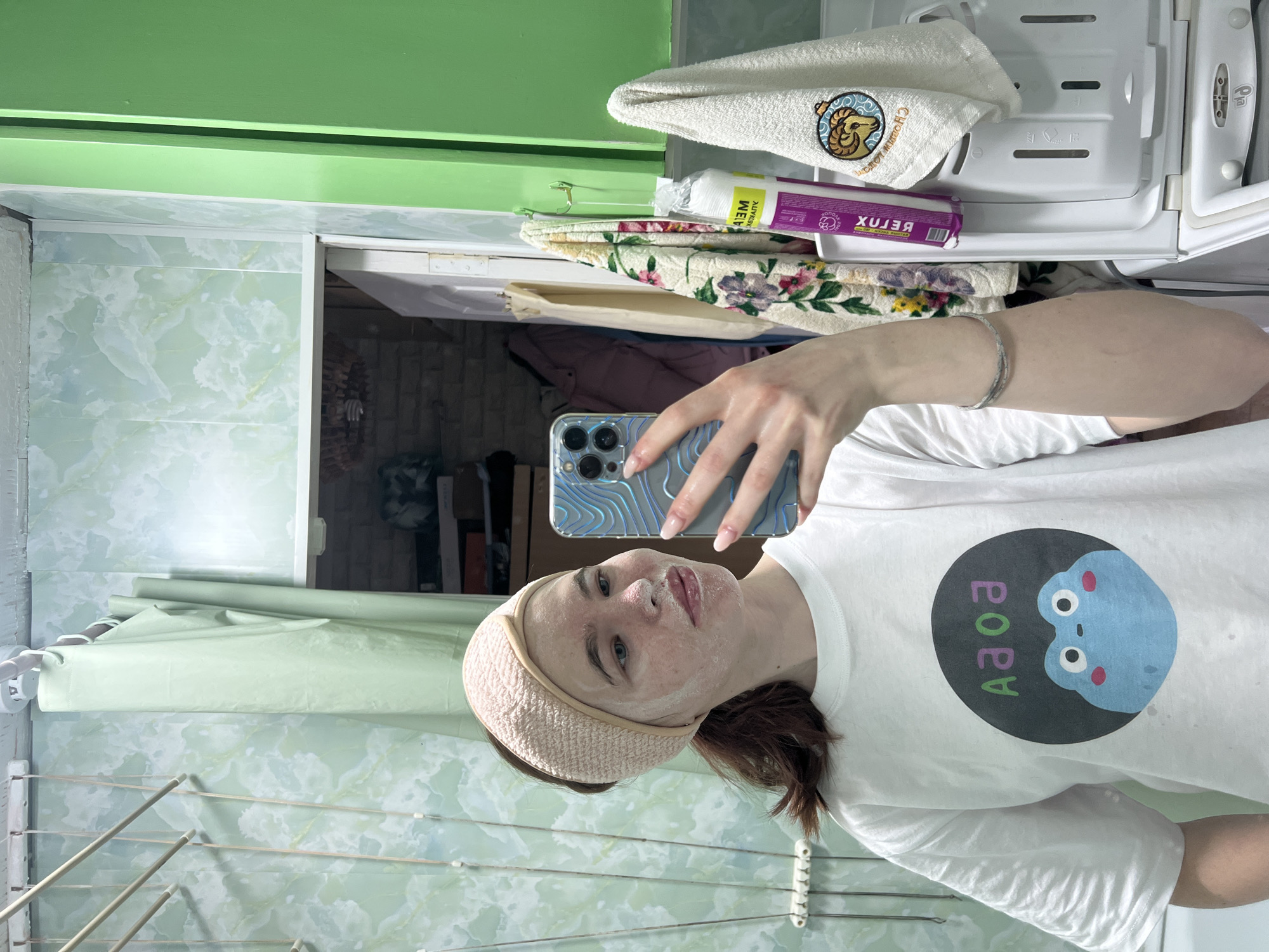 A woman is taking a selfie in a bathroom. She is wearing a white t-shirt with a circular image of a blue animal on it and a light pink headband. Her face has a white substance applied to it. She is holding an iPhone with a light blue patterned case. In the background, there is a green painted wall, patterned tiles, a clothes drying rack, a shelf unit holding towels and personal care products, and a doorway. The location is likely in Chaikovsky, Russia, based on information associated with similar images.