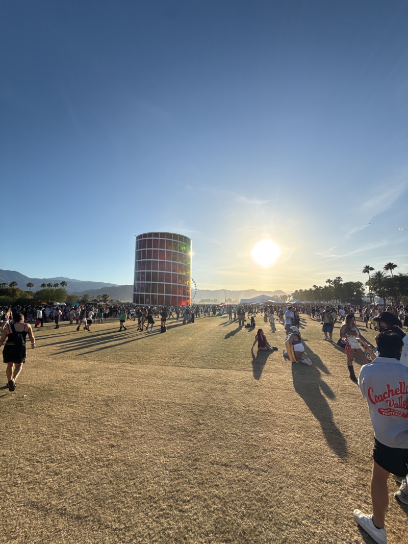 The image depicts a sunny, outdoor scene at the Coachella Valley Music and Arts Festival, located in Indio, United States. A large crowd of people is dispersed across a grassy field, with a cylindrical structure featuring a grid-like design standing prominently in the middle ground. The sun is positioned high in the clear blue sky, creating long shadows across the ground. Palm trees are visible on the right, and mountains can be seen in the background. Some individuals are walking or standing, while others are sitting on the grass. A person wearing a "Coachella Valley Music and Arts Festival" shirt is partially visible in the foreground. The overall atmosphere suggests a lively and crowded event.