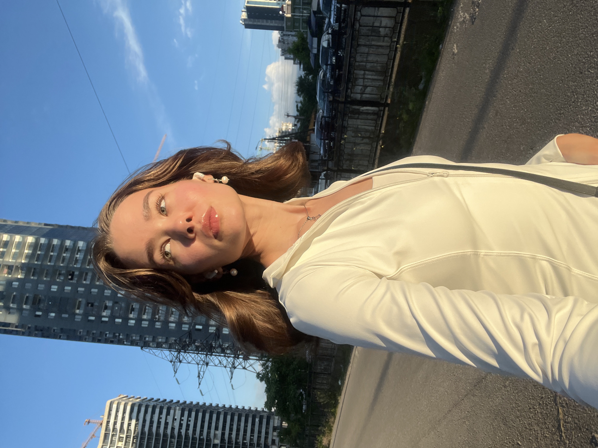 The image shows a young woman with light brown hair and light blue eyes looking upwards and to her right. She is wearing a white zipped jacket over a white top, pearl earrings, and a silver necklace. She has lip gloss on. Her right arm is visible, resting at her side. Behind her, to the left, are two tall buildings under construction, with a crane visible on the left-most building. To the right of the buildings is a structure with numerous wires, likely power lines. Further in the background, there are more buildings and a street with parked cars. The scene appears to be outdoors on a paved surface, possibly a sidewalk or road, with a dark fence bordering the right side of the path. The lighting suggests it is late afternoon or early evening, with the sun casting long shadows. The presence of tall, modern buildings and construction sites is indicative of an urban environment, consistent with the city of Batumi, Georgia, known for its rapid development and skyline dominated by high-rise structures.