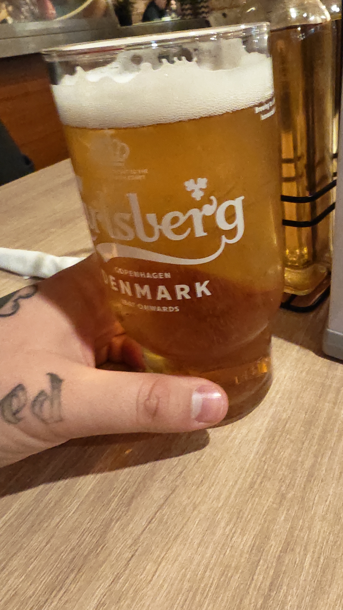 The image shows a partially consumed pint of Carlsberg beer in a glass, held by a hand with tattoos. The beer has a frothy white head. A bottle with a golden liquid is visible in the background. The scene is set on a light-colored wooden table. There is no information in the image about the location in the city of Plovdiv, Bulgaria.
