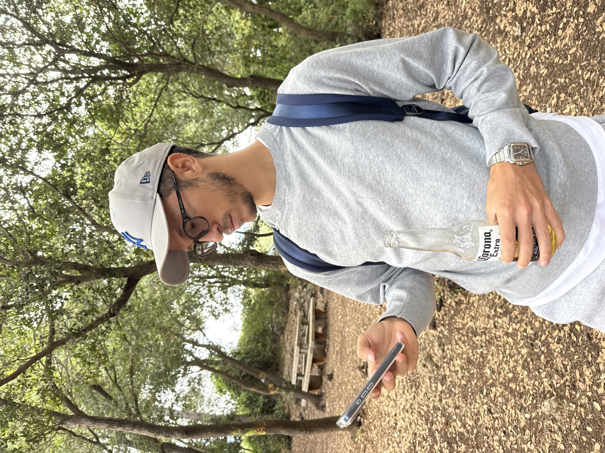 A man stands in a wooded area, likely a park, location unspecified, although presumed to be near Nesher, Israel. He is wearing a grey sweatshirt, grey baseball cap with a blue logo, glasses, and a silver watch. He is wearing a backpack. He holds a cell phone in his left hand and a bottle of Corona Extra beer in his right. Fallen leaves cover the ground. A picnic table is visible in the background. The lighting suggests a sunny day.