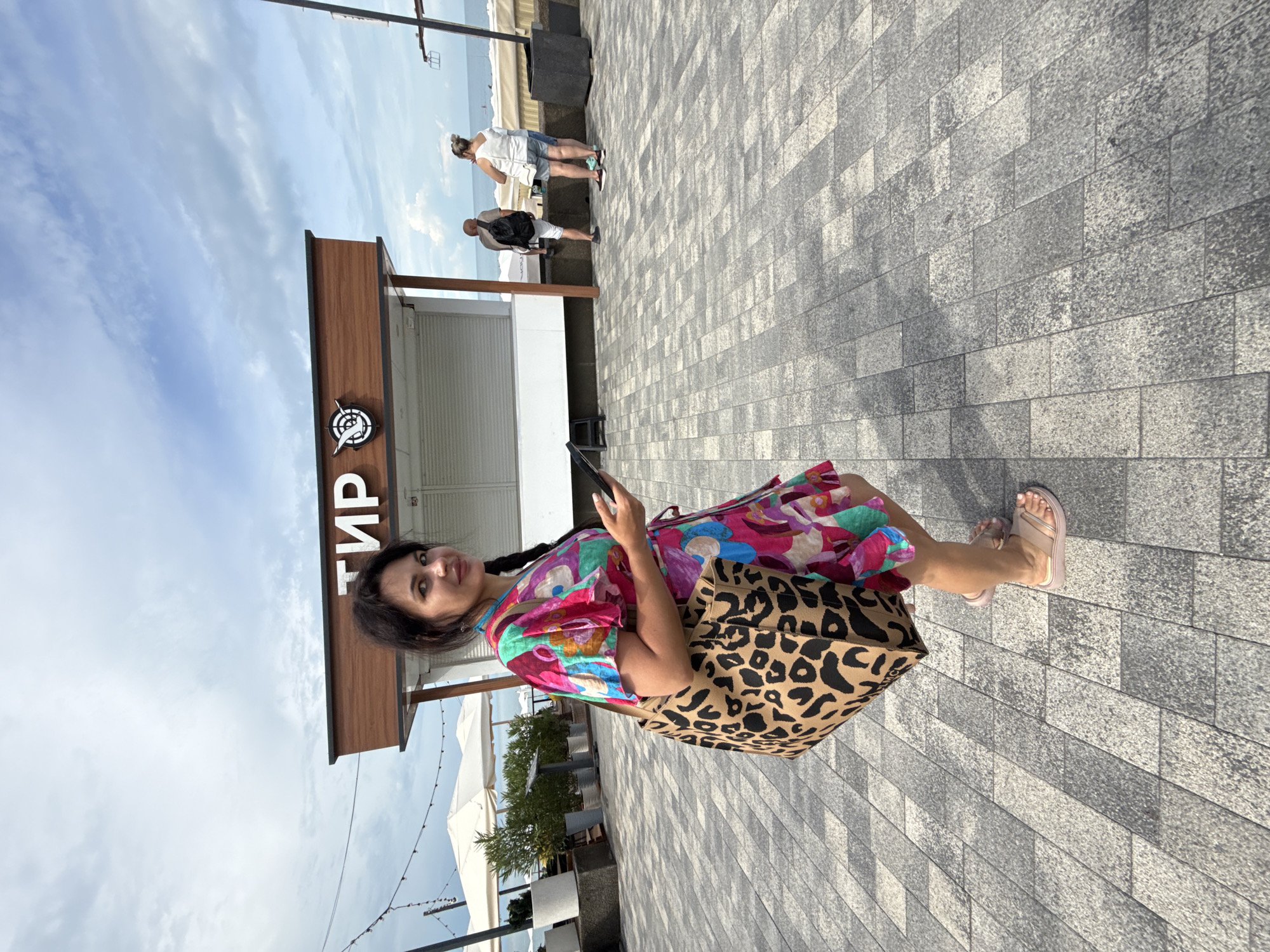 A woman with dark hair and a braid is walking on a paved promenade in Sochi, Russia, holding a black mobile phone in her right hand. She is wearing a colorful, patterned dress with short sleeves and a leopard-print tote bag slung over her left shoulder. Her feet are clad in light-colored slides. Behind her, a food kiosk with the Cyrillic letters "ТИР" is visible. In the background, two people are standing near the sea, and there are white umbrellas and greenery to the left. The sky is partly cloudy.