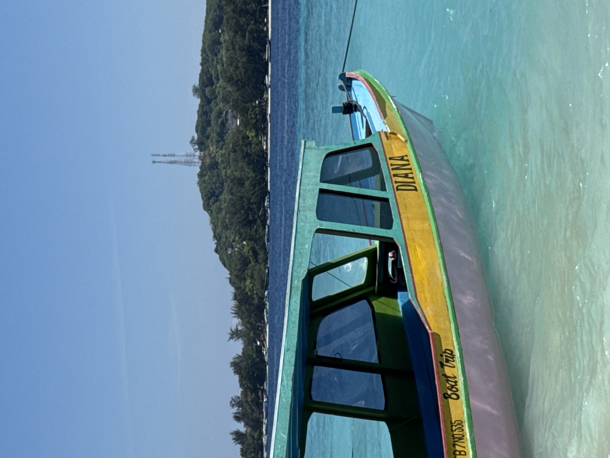 A colorful wooden boat named "DIANA" is depicted in calm, clear turquoise waters under a bright, cloudless blue sky. The boat, possibly a traditional Indonesian vessel, is painted with vibrant blue, green, and yellow trim, and a lighter grey or purple hull. Windows are visible along its side. A mooring rope extends from the bow, suggesting it is anchored or docked.
The boat is positioned in shallow, crystalline water, which transitions from a very light, almost white-blue near the boat to a deeper blue further out, indicative of varying depths. On the left side of the frame, a lush green island covered with dense tropical vegetation stretches horizontally. Two tall, slender metal structures, resembling communication towers, rise above the tree line on the island.
The scene conveys a serene tropical setting on a sunny day. The visible text on the boat reads "DIANA" in yellow lettering, "Boat Trip" in black below it, and "B 7NO,535" in white lettering near the hull. No people are visible, and the boat appears to be idle, perhaps awaiting passengers or moored after an activity. The overall impression is one of a peaceful island getaway or a starting point for a boat excursion.