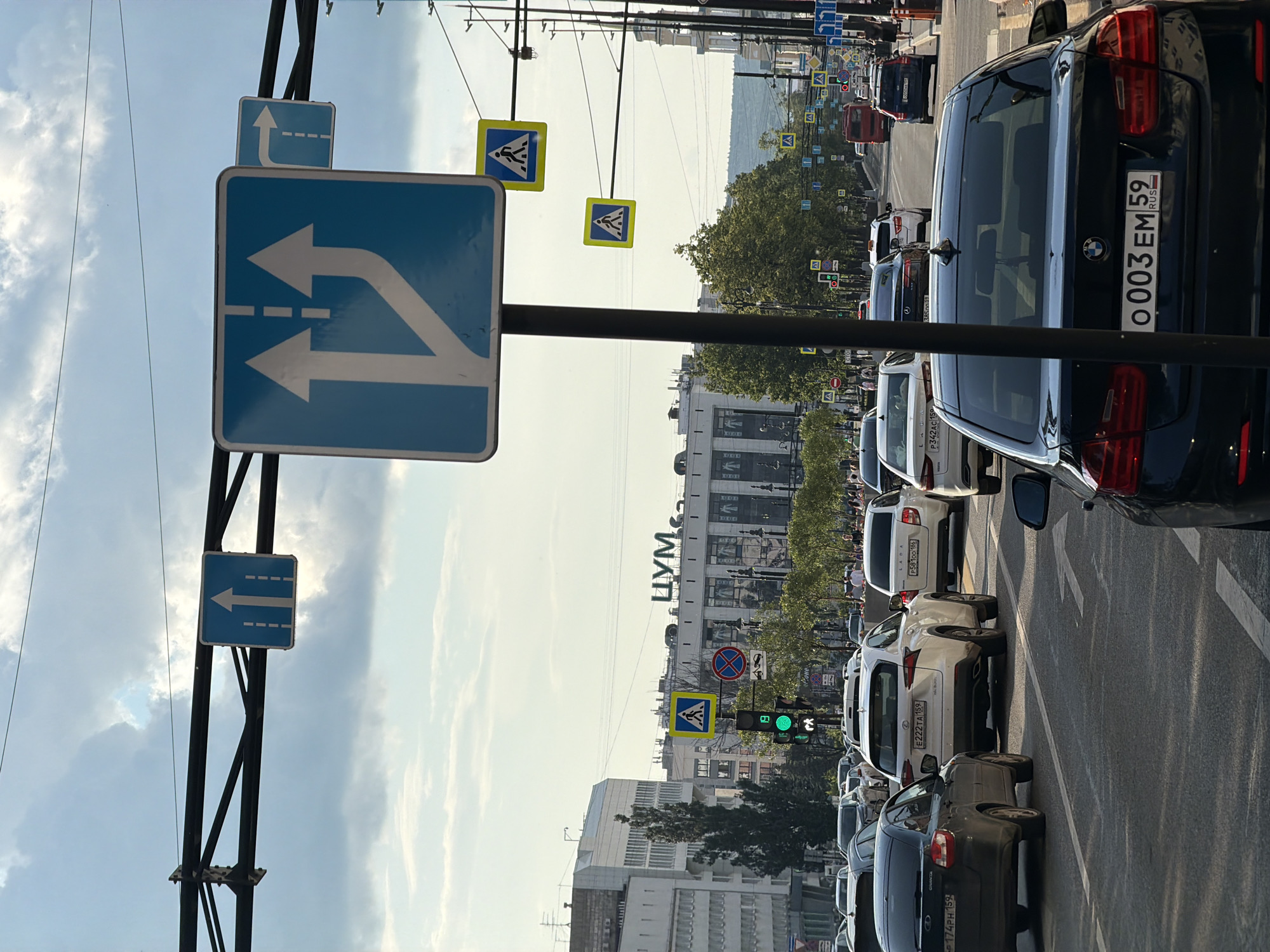 The image captures a traffic scene on a sunny day in Perm, Russia. The foreground shows the rear of a dark BMW sedan, license plate "o003EM 59 RUS", on a multi-lane road. Ahead, a white Lexus SUV and other vehicles are visible. Traffic lights are green, and road markings indicate multiple lanes. Overhead, a complex structure of wires and traffic signs is suspended. One prominent blue sign depicts two arrows pointing upwards, with one lane splitting to the right, indicating lane management. Another blue sign shows a single arrow turning right. Yellow pedestrian crossing signs are also visible. In the background, several multi-story buildings line the street, one bearing the Cyrillic signage "ЦУМ". Trees and pedestrians are present along the sidewalks. The sky is partly cloudy, with a few dark clouds visible against a lighter sky. The overall impression is of a busy urban intersection in Perm.
