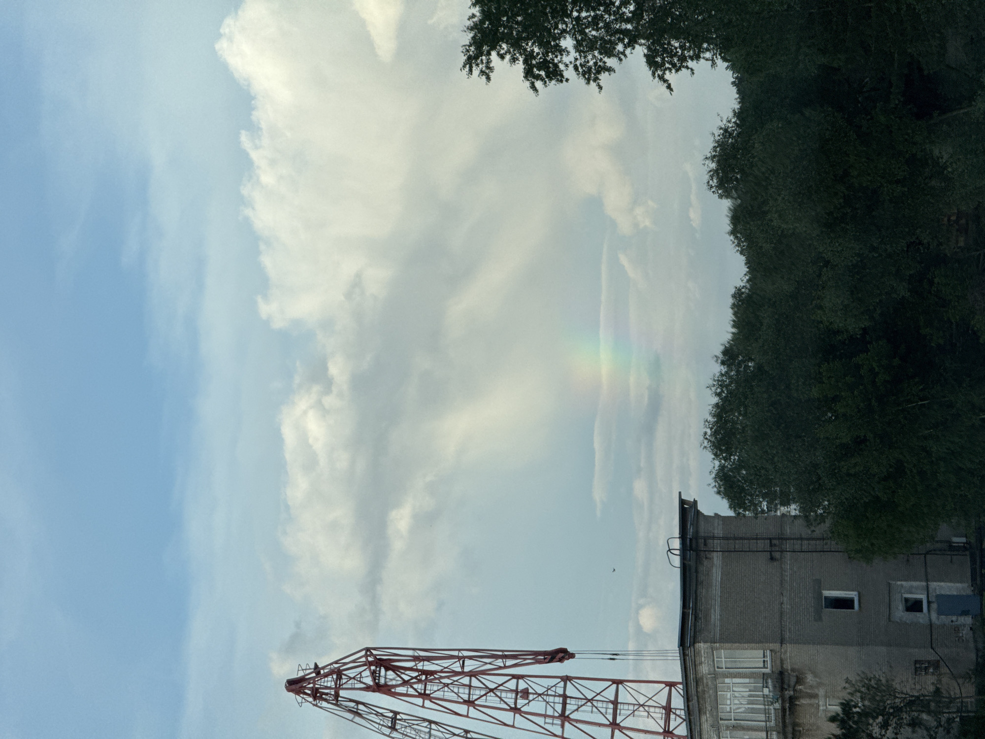 The image shows a sky with light blue and white clouds. A faint rainbow is visible among the clouds. A portion of a red crane is visible in the lower left corner. To the right is a gray concrete building with windows and a ladder on the side. Trees are at the top of the image. The location is in the city of Chaikovsky, Russia. There are no people visible. A small bird is flying in the sky between the building and the crane.