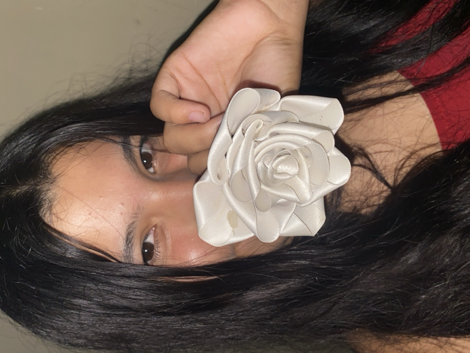 A person with dark hair is holding a cream-colored, artificial rose in front of their face. The flower is made of ribbon and covers part of their cheek. The person's eyes are visible, and they appear to be looking at the camera. The lighting is dim, and the background is blurred. The individual is wearing a red top.
