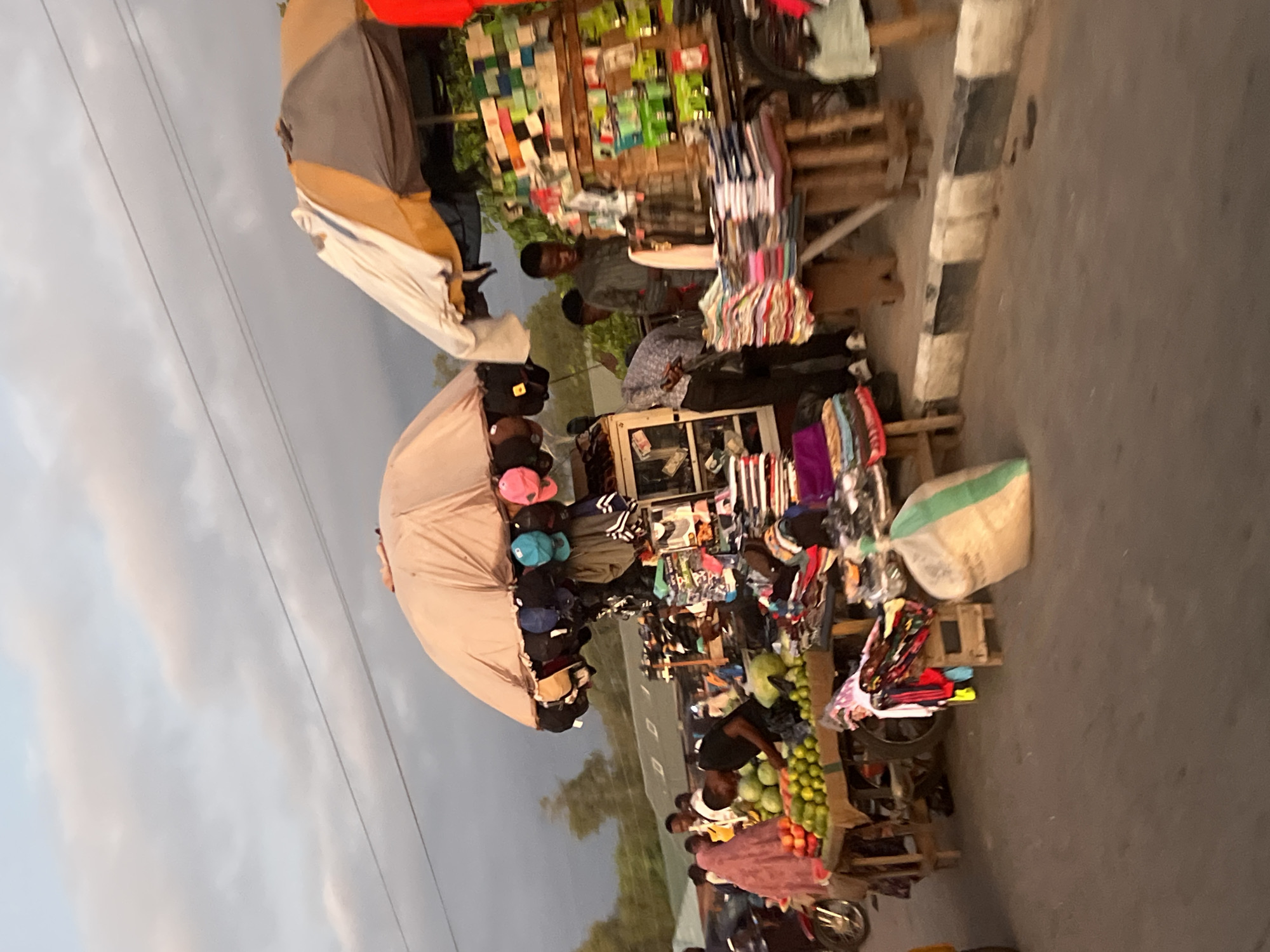 This image depicts an outdoor street market scene in Howayi, Nigeria, occurring under an overcast sky, likely during late afternoon or early evening. The foreground features a paved road with a black and white striped curb on the right. Several market stalls line the road, each protected by large umbrella canopies.
On the left, a stall displays an abundance of fruits, including red apples, green apples, and what appear to be oranges or limes, arranged on a wooden table. A woman dressed in a pink or maroon hijab and dress stands in front of this stall, looking at the produce. Behind the stall, a man in a black shirt is seen attending to the fruits. A portion of a motorcycle is visible on the far left, parked near the curb.
The central stall is covered by a large, light brown umbrella from which numerous baseball caps in various colors (blue, pink, black, beige) are suspended for display. Below the umbrella, a wooden table holds stacks of folded clothing and other merchandise. To the left of this table, a display cabinet shows additional items. A large, transparent bag containing a light-colored substance, with green markings, rests on the ground in front of this stall. A man in a light-colored, patterned shirt is partially visible behind this stall.
To the right, another stall features a multi-colored umbrella (white, brown, beige). This stall has a wooden tiered display unit filled with various packaged goods, possibly small electronics or household items. Below, stacks of folded garments are neatly arranged on a table. A man in a dark grey or blue shirt stands behind this stall, observing the merchandise. Other individuals are present in the background, moving around the market. Overhead power lines traverse the sky in the upper portion of the image.