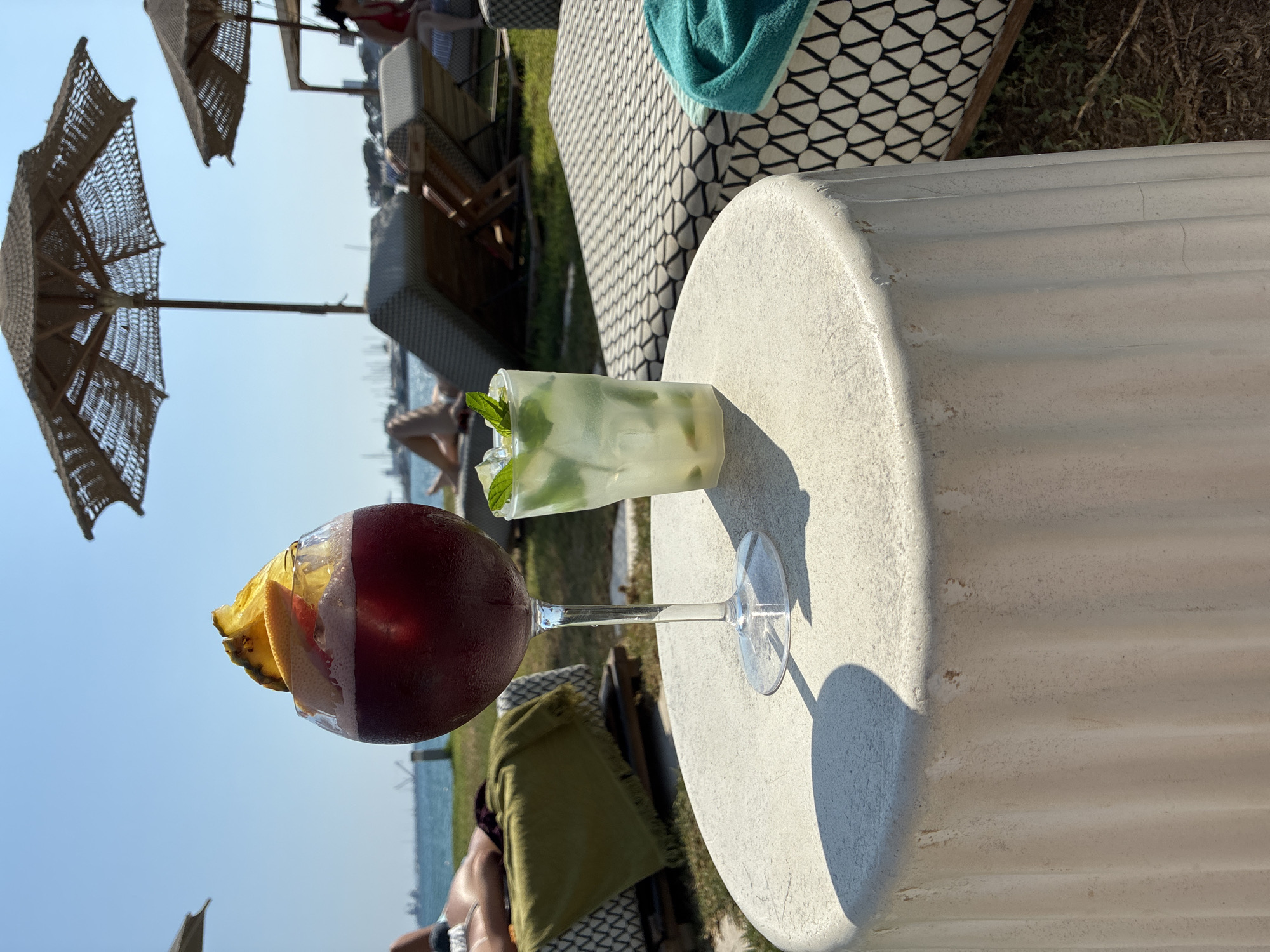 Two drinks are placed on a white textured cylindrical table at a beachfront resort in Limassol, Cyprus. The drink on the left is in a large wine glass and is a dark red color, possibly sangria or a similar wine-based cocktail, garnished with a slice of pineapple and orange. To its right is a clear glass filled with a light-colored drink, likely a mojito, with ice, mint leaves, and lime. In the background, people are relaxing on lounge chairs under large woven umbrellas on a grassy area overlooking the Mediterranean Sea. The sky is clear and blue.