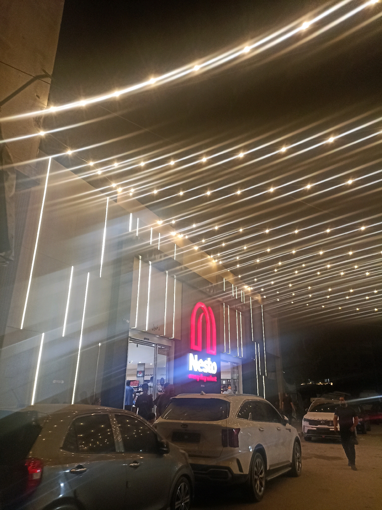 This image depicts the entrance of a "Nesto" hypermarket at night in Gaza City, Palestinian Territories. The building features a modern facade with numerous horizontal strings of warm-toned lights, creating a visually striking illuminated canopy. Vertical white LED strips accent the building's exterior. A prominent red neon sign displaying "Nesto" is visible above the entrance.
In the foreground, several cars are parked. A gray car is closest to the viewer, angled towards the right. To its right is a white SUV, and further back, another white SUV or car is partially visible. Several individuals are seen entering and exiting the hypermarket through the automatic glass doors. One person in dark clothing stands near the white SUV on the right, while others are inside the store or walking nearby. The ground appears to be a dirt or gravel surface. The overall atmosphere is illuminated by the building's lighting and the ambient nighttime sky.