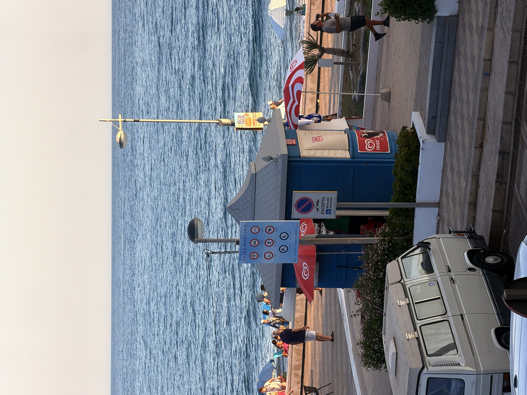 The image depicts a scene along a seaside promenade in Castillo Bajo-conejito, Spain. The sky is a pale, cloudless hue, transitioning to a calm, blue ocean. A paved promenade runs parallel to the sea, with a white-tiled wall separating it from a grassy area.
On the promenade, several people are visible. Some are seated on beach chairs or towels near the water's edge, partially obscured by blue umbrellas. Others stand or walk along the promenade. A man in a dark blue shirt and white shorts stands facing the sea, while another man in a grey t-shirt and dark shorts walks away from the camera, holding a water bottle.
A prominent blue kiosk with a grey roof is situated on the promenade. It has signs and awnings. One sign, a large blue rectangular placard, displays multiple circular icons with red borders, indicating regulations for the promenade. Another smaller sign below it shows a red circle with a diagonal line over a wheelchair symbol, and text that reads "EXCEPTO MOVILIDAD REDUCIDA." A red and white striped umbrella is positioned in front of the kiosk. Two red Frigo ice cream logos are visible on the kiosk's side.
To the left of the kiosk, a white camper van is parked. It has several windows and a raised roof section. A blue wheelchair symbol is affixed to one of the van's windows.
Further down the promenade, a lamppost stands near the kiosk. A small yellow sign is attached to the lamppost with the text "FOOD TRUCK ESPINOS TORREX." Another, larger white umbrella with a blue canopy is visible in the distance near the water.
The ground is paved with interlocking diamond-shaped tiles. In the foreground, a section of a road or lower pathway is visible with a metal grate. Lush green bushes and small pink flowers line parts of the promenade, particularly near the kiosk and the camper van. The overall impression is a public space by the sea, likely during daytime hours, with people enjoying the coastal environment.