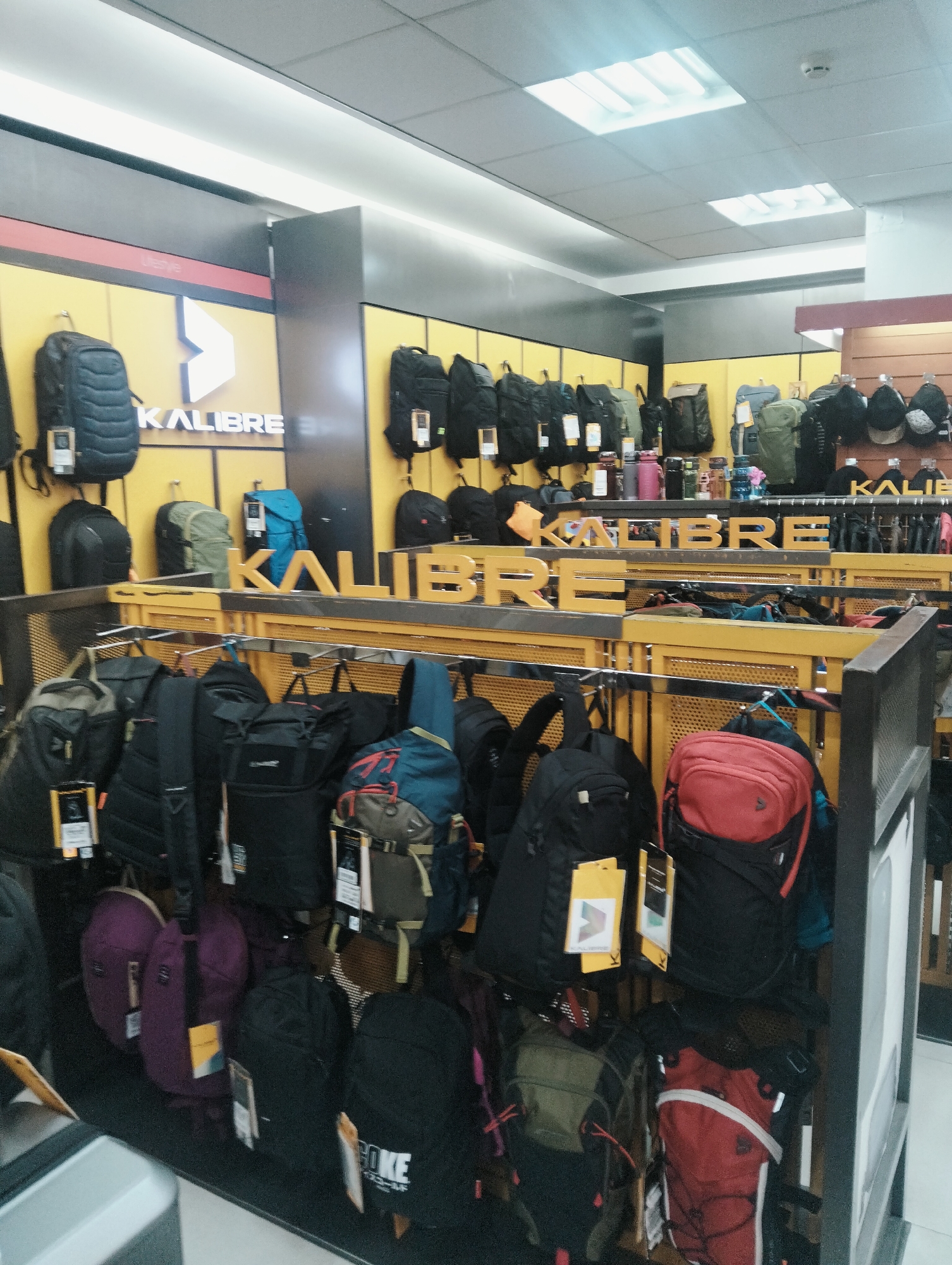 The image shows a retail store interior, likely a department store or specialized outdoor goods shop, in the city of Medan, Indonesia. The store is well-lit with overhead fluorescent lighting. The dominant display features a variety of backpacks of different sizes, colors, and brands, primarily Kalibre. Backpacks are hung on racks and shelves, and some are displayed on the floor. The walls are decorated with yellow panels and signage bearing the Kalibre logo. On the shelves, there are also water bottles and caps. The overall environment is clean and organized, suggesting a commercial setting focused on selling travel and outdoor equipment. There are no people visible in the image.