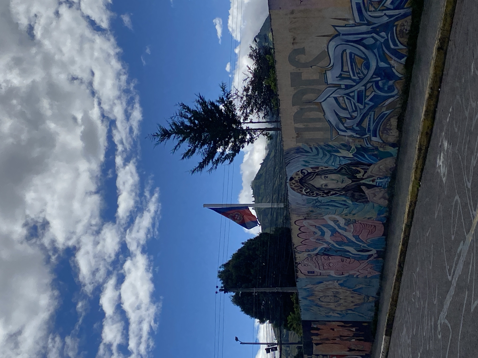 The image shows a view in Quito, Ecuador. A wall mural with graffiti is on the right. A flag of Quito is flying in the wind in the center of the image. Trees and a mountain are in the background, behind the flag. The sky is blue with white clouds. A street is on the bottom right.