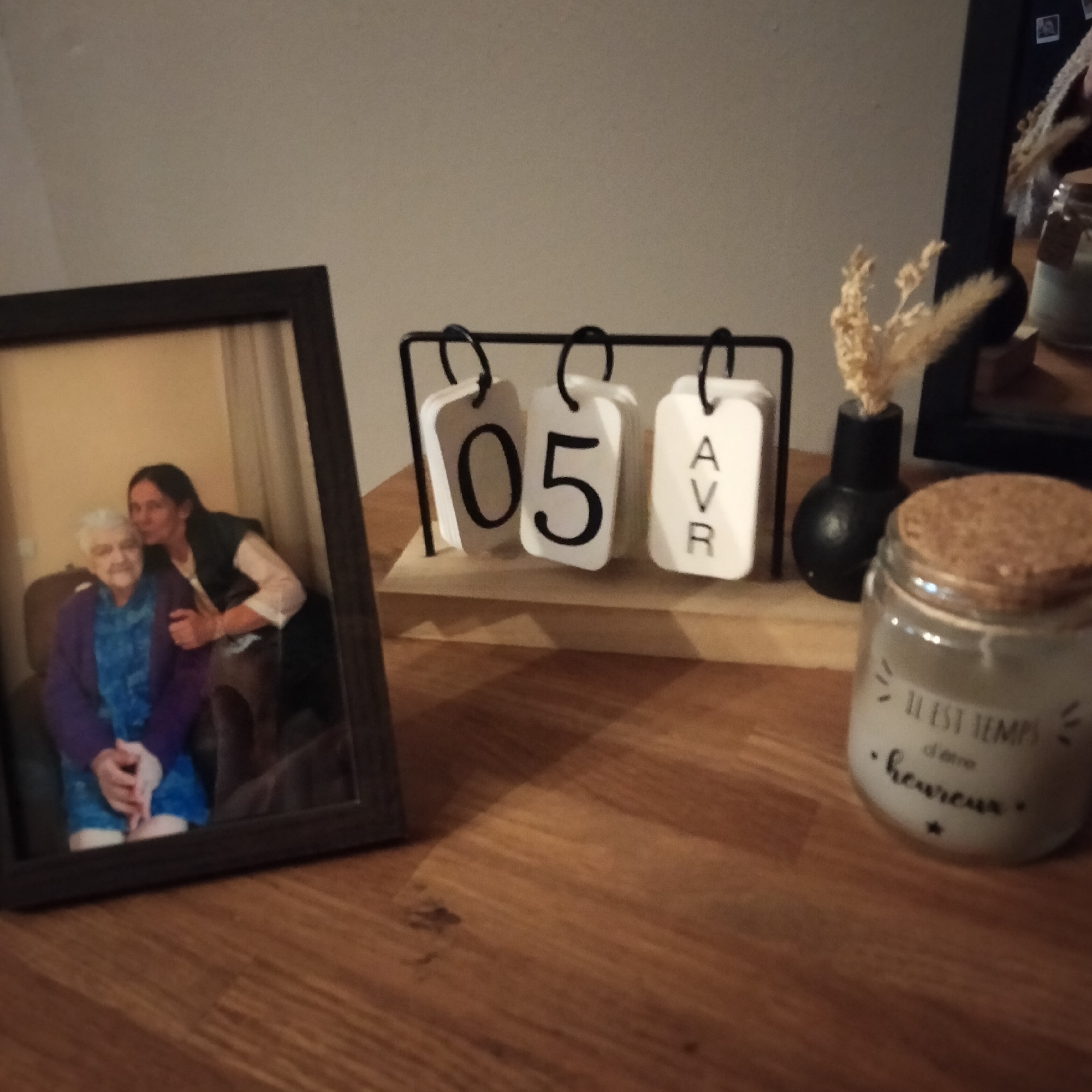 A photograph shows a wooden desk or tabletop. On the left, there is a framed photograph of an older woman seated with a younger woman embracing her. Center in the frame sits a perpetual calendar displaying "05 AVR," indicating April 5th. To the right of the calendar, there is a small black vase with dried flowers and a candle in a jar with a cork lid. The jar has French text on it, "IL EST TEMPS d'être heureux." At the background, there is a dark frame. The scene appears to be indoors, in Lannoy, France.
