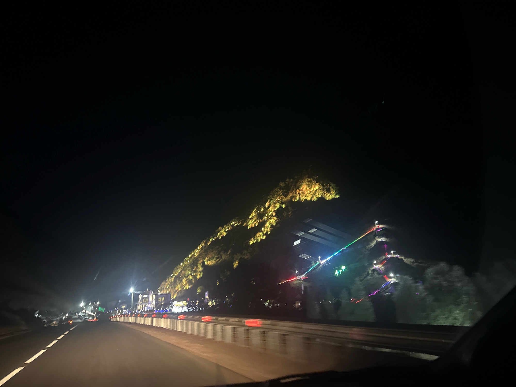 The image depicts a nighttime view from a vehicle traveling on a road in Arashan, Kyrgyzstan. The road is paved with white lane markings visible. To the right of the road, a series of illuminated structures and natural terrain are visible. A hillside is adorned with numerous golden lights, creating a shimmering effect. Further along the hillside, a path or staircase is illuminated with colorful lights, including green, blue, purple, and red. Several buildings with lights are also visible in the background. The sky is dark and clear. The overall scene suggests a festive or illuminated tourist area within the city of Arashan.