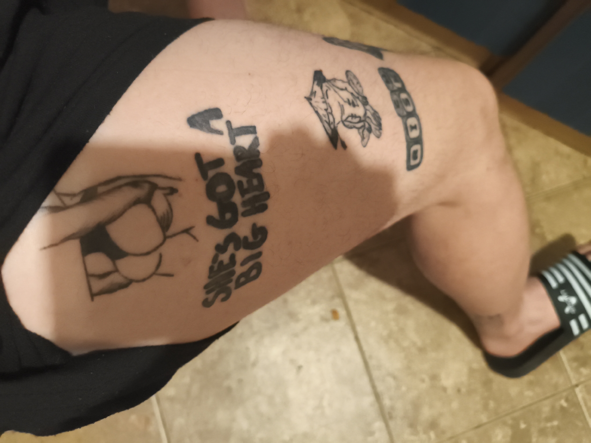 A view from above shows a person's left leg with multiple black ink tattoos. The person is wearing black shorts and a black and white striped sandal on their left foot. The tattoos include what appears to be the outline of buttocks, the phrase "SHE'S GOT A BIG HEART," a sketched cow head, and a block of shapes. The leg is positioned on a light brown tiled floor. The location is not discernible from the image.