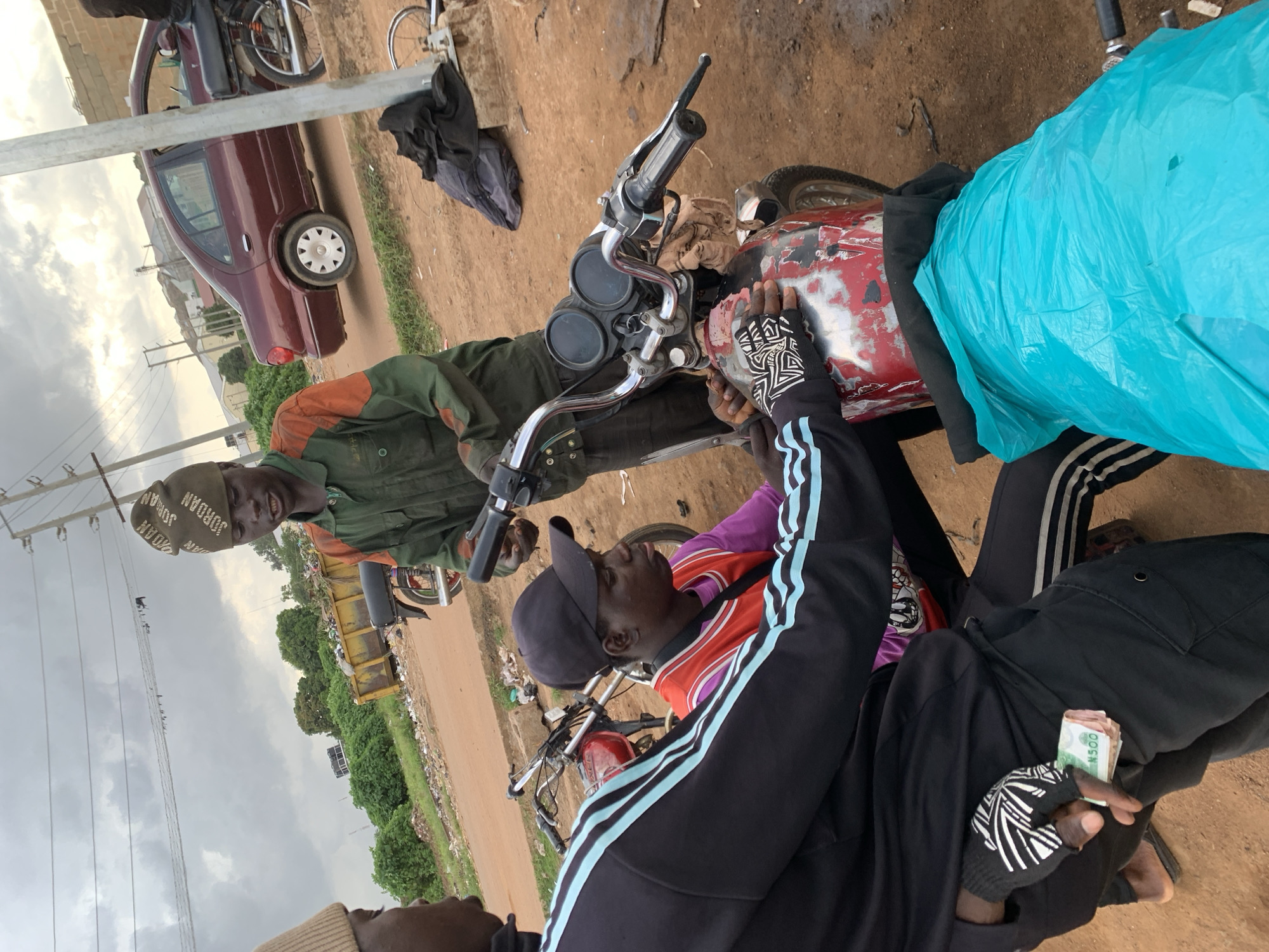 In Barakin Lerawi, Nigeria, three young men are gathered around a red motorcycle that appears to be undergoing repairs or modification. The man on the far right, wearing a green and orange work jumpsuit and a "Jordan" branded beanie, is actively working on the motorcycle's handlebars. To his left, another young man in a purple shirt and red vest with a black baseball cap is also focused on the motorcycle. In the foreground, a third young man, partially obscured, wears a black tracksuit with blue stripes and fingerless gloves. He is holding a wad of Nigerian currency, seemingly N500 notes, tucked into his pocket. The environment is dusty and appears to be an outdoor workshop or informal gathering space, with a pile of trash in a yellow skip and a dark red sedan parked in the background. The sky is overcast.