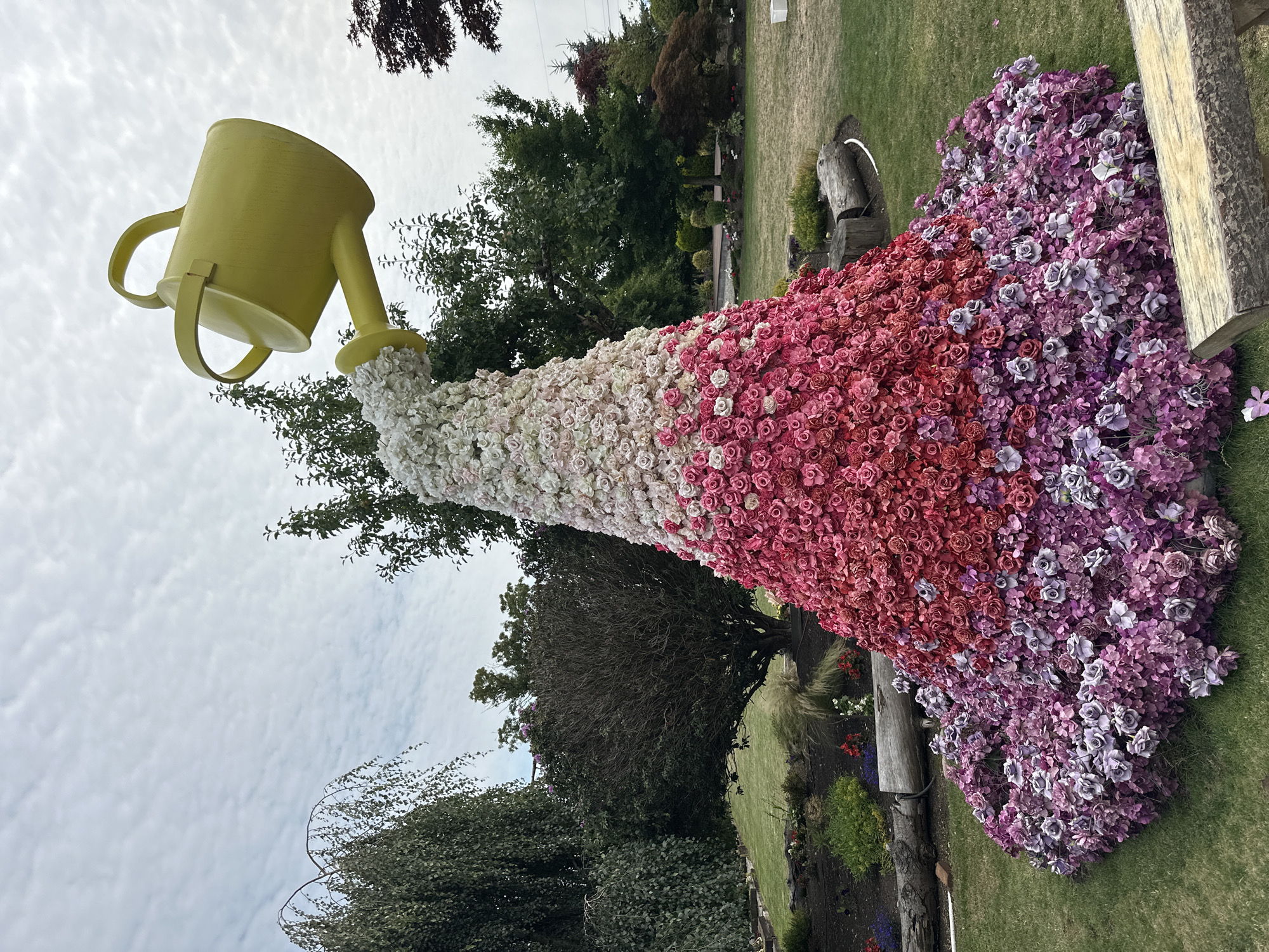 An outdoor floral sculpture resembling a watering can pouring a cascade of flowers is situated in a garden setting. The sculpture is conical in shape, starting with clusters of purple and pink flowers at the base, transitioning to red and pink flowers in the middle, and culminating in white flowers at the top. A large, bright yellow watering can is positioned above the sculpture, tilted as if pouring the flowers. The sculpture is surrounded by green grass, trees, and some landscaping. The sky is overcast with scattered clouds. This sculpture is located at The Gardens at Queen Elizabeth Park in Richmond, Canada.