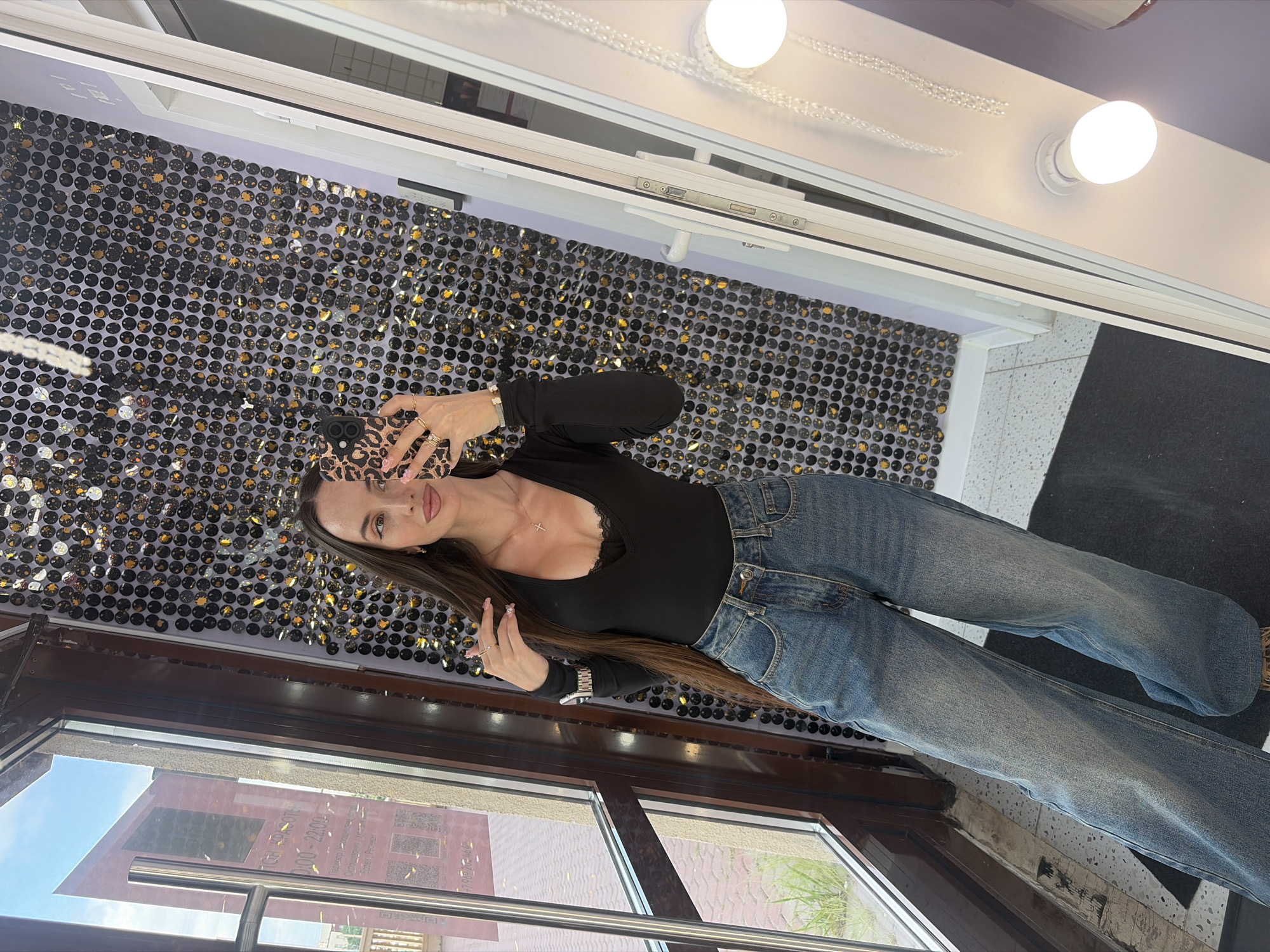 A young woman with long brown hair and fair skin is taking a mirror selfie in what appears to be a salon or retail establishment in Saint Petersburg, Russia, as indicated by the Cyrillic text visible on a sign through a large window. She is wearing a long-sleeved black top with a lace trim at the V-neckline and a delicate silver cross necklace. Her bottom half is adorned with a pair of light blue, distressed, wide-leg jeans. She is holding a leopard-print phone case in her right hand, angled to capture her reflection. Her left hand is casually resting on her head, near her hair. The background is a wall covered in shimmering black and gold sequins, creating a striking visual effect. To her left, a large window offers a view of the exterior, revealing a building with red signage. To her right, a white doorframe with a light fixture featuring multiple bulbs is visible, suggesting a dressing or makeup area. The floor is a combination of dark matting and speckled tile. The overall impression is one of a trendy and stylish environment.