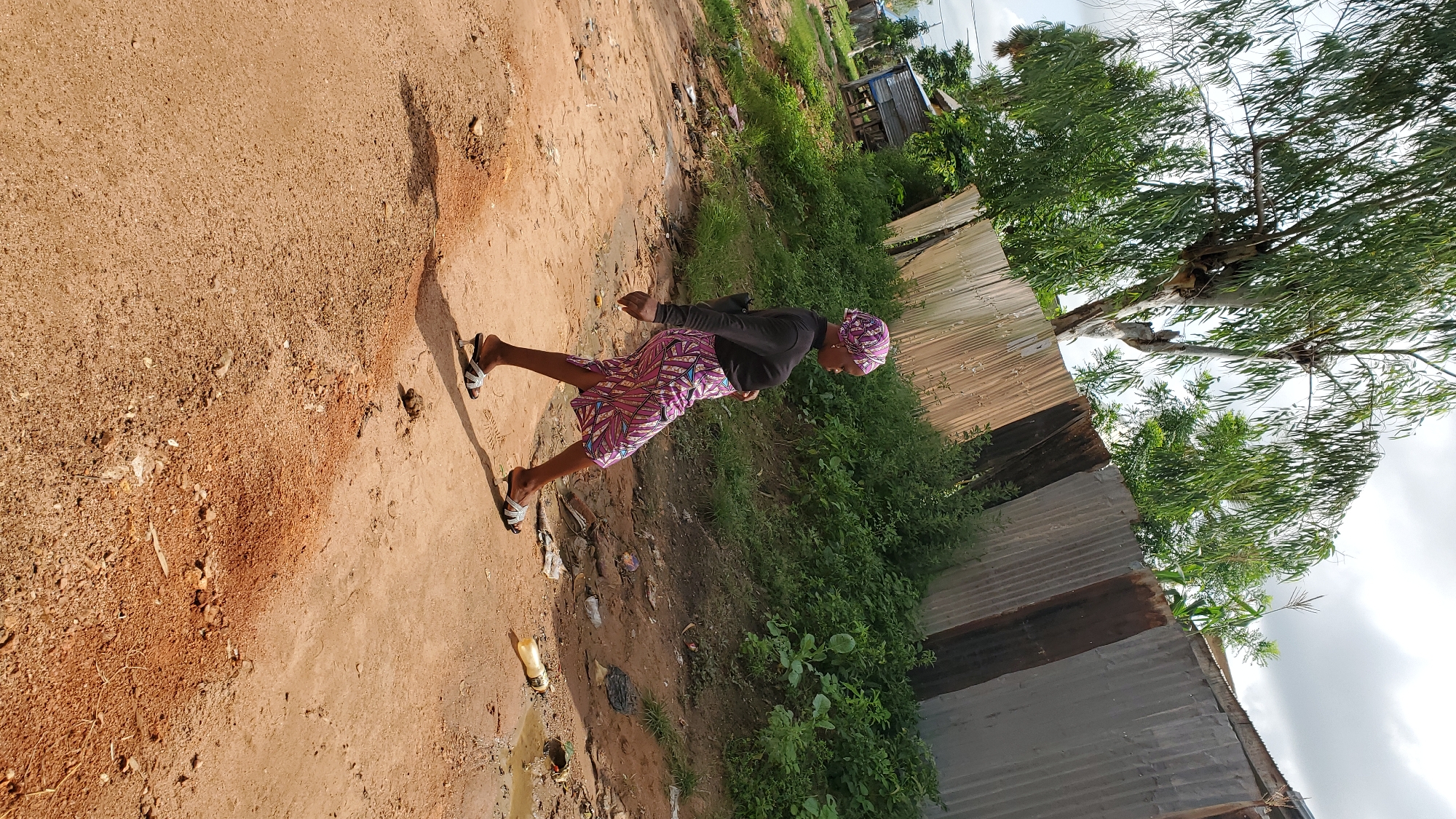A full-shot, eye-level, medium-angle, outdoor, daytime, slightly tilted image shows a woman walking on a dirt road in Jalingo, Nigeria. The woman is wearing a black long-sleeved shirt, a colorful patterned skirt, and sandals with clear heels and white straps. She has a headscarf that matches her skirt. She is walking towards the right of the frame, with her body angled in a walking pose and her head turned to the left. She is carrying a black bag over her left shoulder.
The road is made of red-brown dirt with scattered rocks and some debris, including a plastic bottle and what appears to be a piece of white plastic. To the right of the road, there is a muddy puddle. Behind the road, there is a slope covered with green vegetation and some taller plants. A fence made of corrugated metal sheets runs along the top of the slope. Beyond the fence, trees with green leaves are visible, as well as a cloudy sky. To the far left of the frame, a small, simple structure with a metal roof is visible in the distance. The lighting suggests it is a sunny day, casting a shadow of the woman on the road.