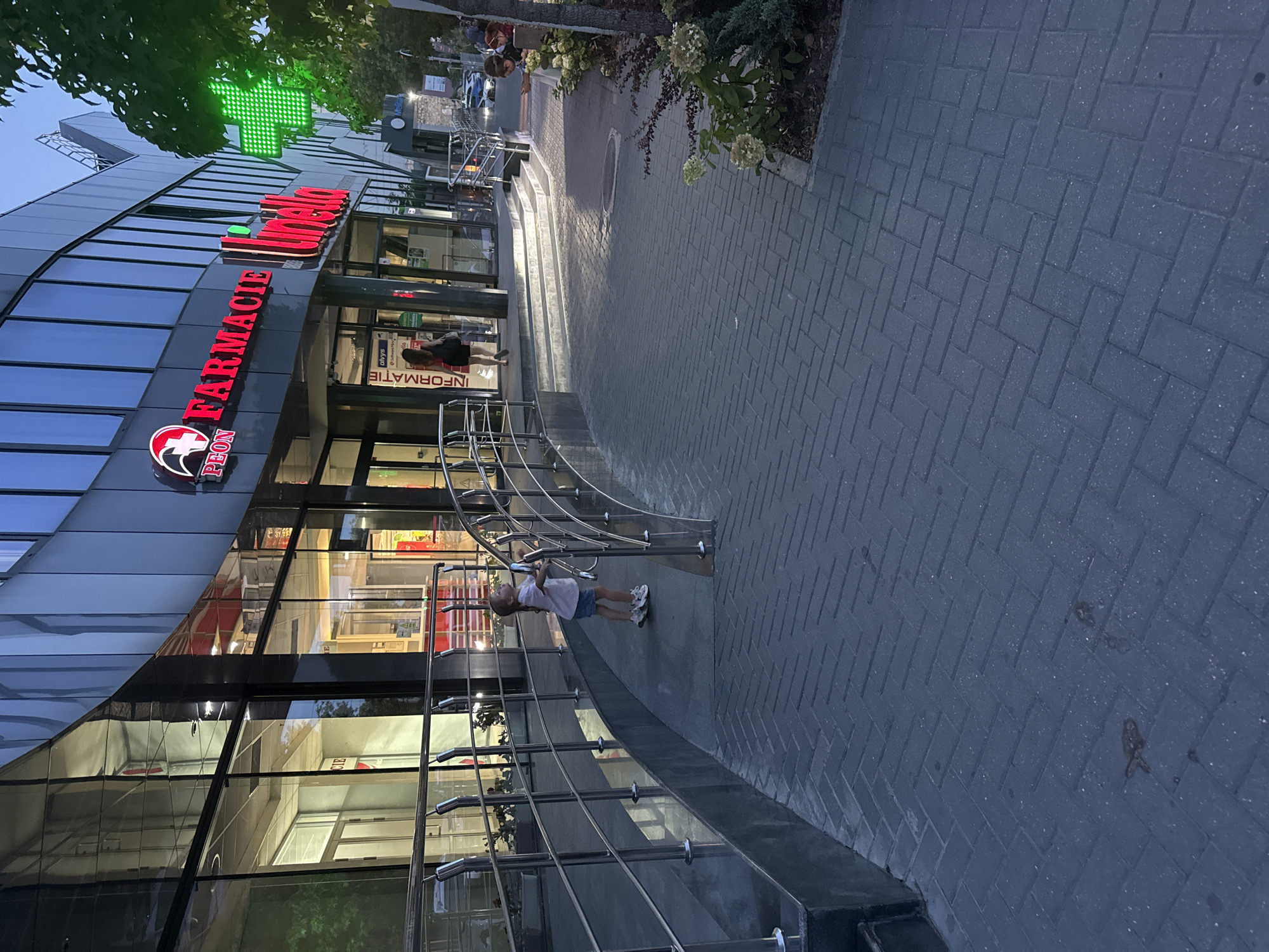A young girl, wearing a light-colored t-shirt and denim shorts, stands on a paved walkway in front of a modern building. She is looking upwards and slightly to her left, her hands resting on a metal railing. The building features large glass windows and a prominent sign that reads "PEON FARMACIE Linella" in red and white lettering. A bright green illuminated cross, a common symbol for pharmacies, is visible to the right of the building, partially obscured by tree foliage. The walkway is constructed of dark gray interlocking pavers arranged in a herringbone pattern. In the background, several cars are parked on a street, and more people are visible near the entrance of the building. The lighting suggests it is dusk. This scene is located outside a Linella supermarket and pharmacy on Calea Ieșilor in Chișinău, Moldova.